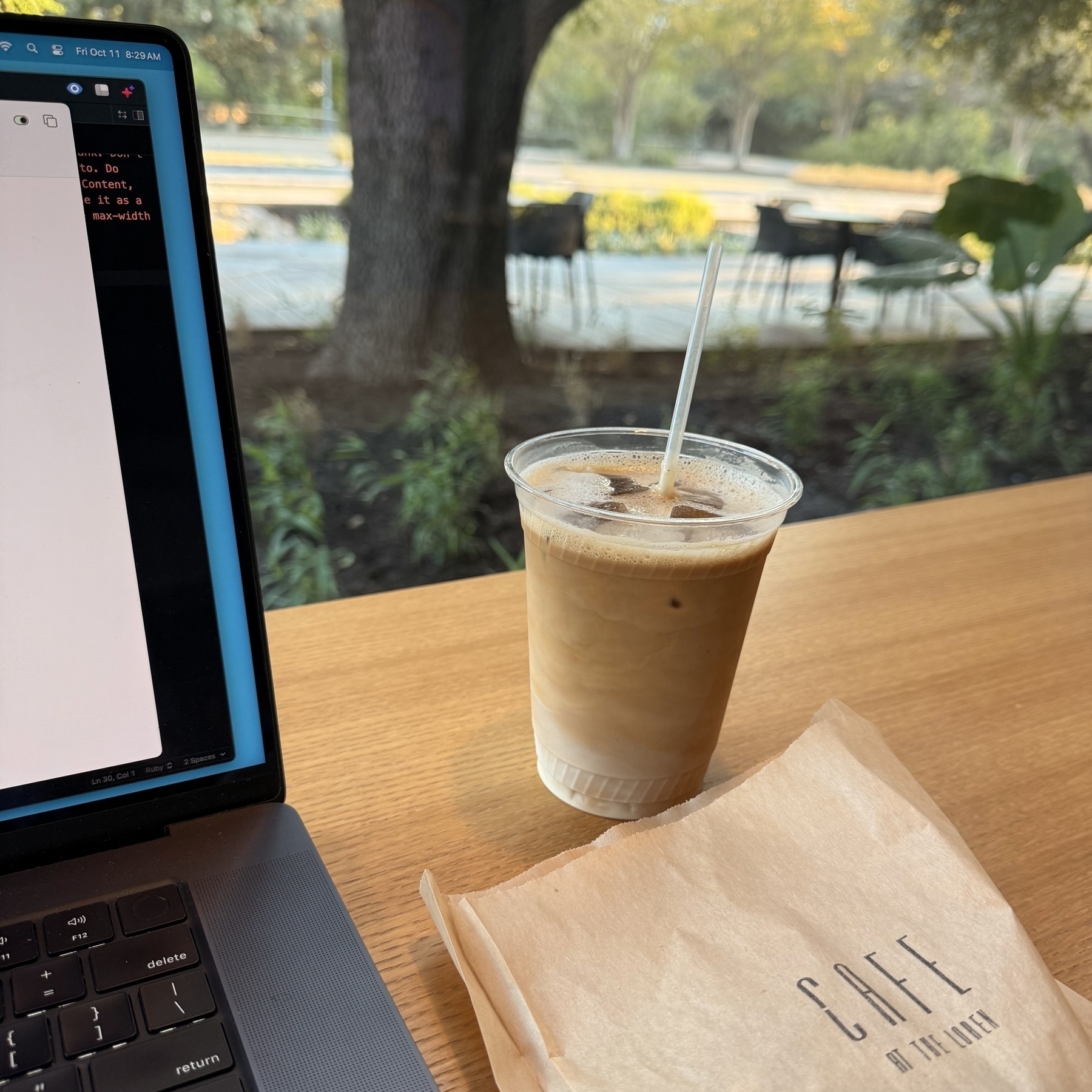 A laptop is on a table next to an iced coffee and a paper bag with probably breakfast inside, and a view of a tree and chairs outside.