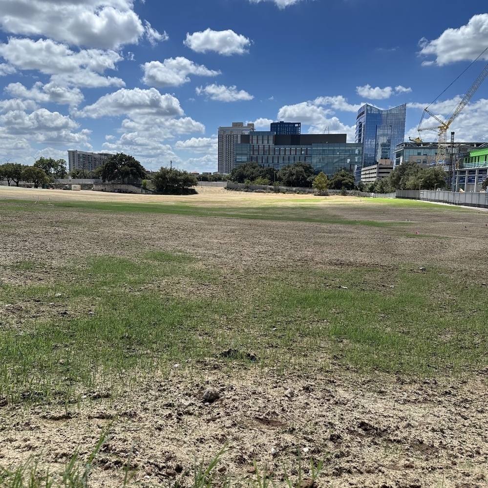 An empty field with patches of grass in front of a group of modern buildings in the distance and cranes under a partly cloudy sky.