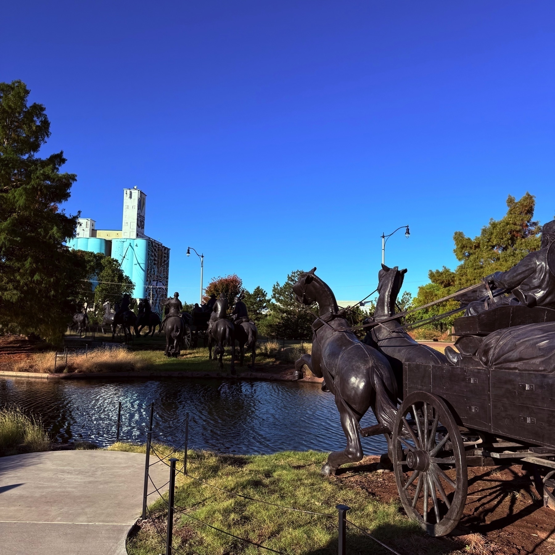 A bronze statue of horses pulling a wagon is set against a backdrop of trees and a small canal, with tall buildings and a clear blue sky in the distance.