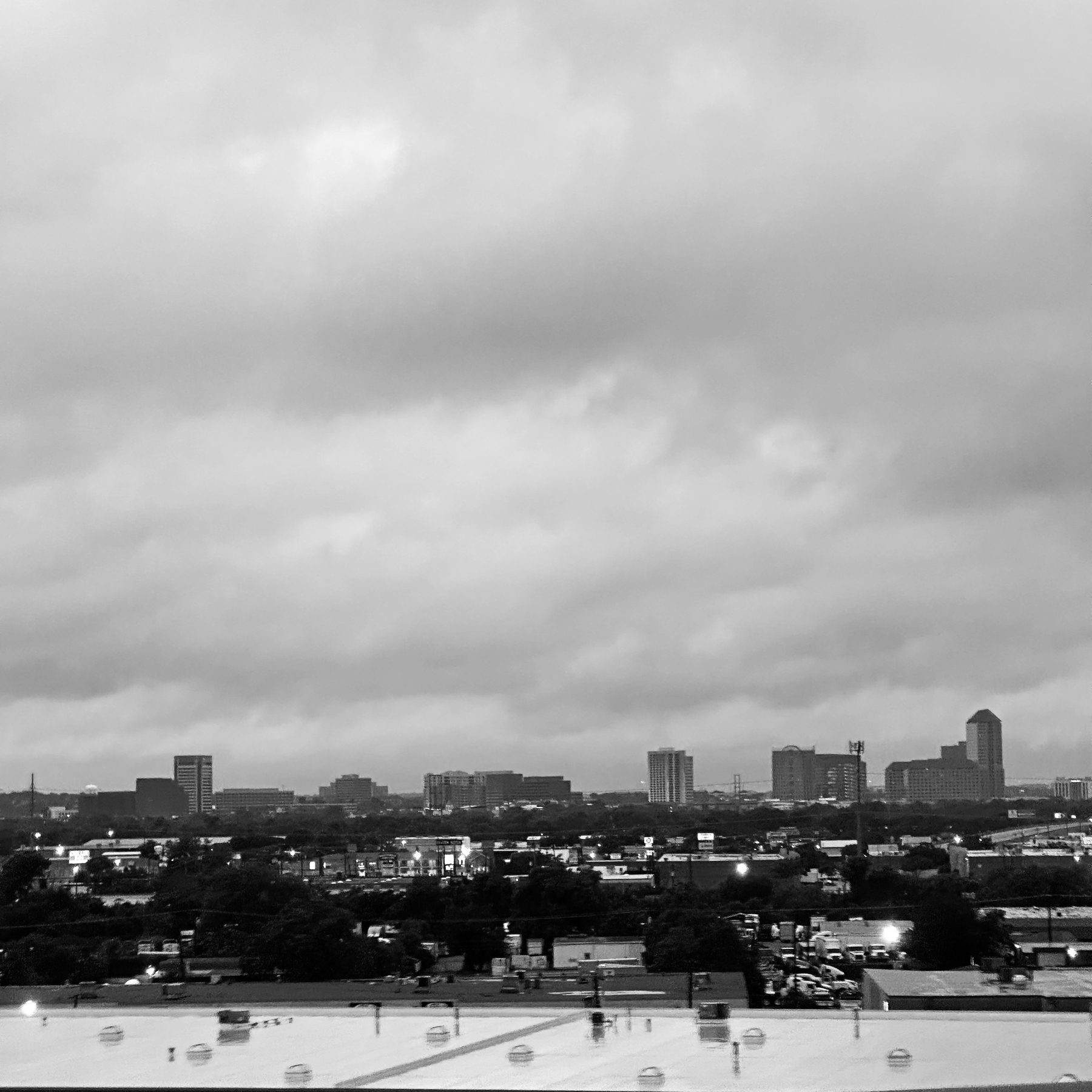 A grayscale cityscape is depicted under a cloudy sky, with buildings silhouetted on the horizon and a rooftop in the foreground.