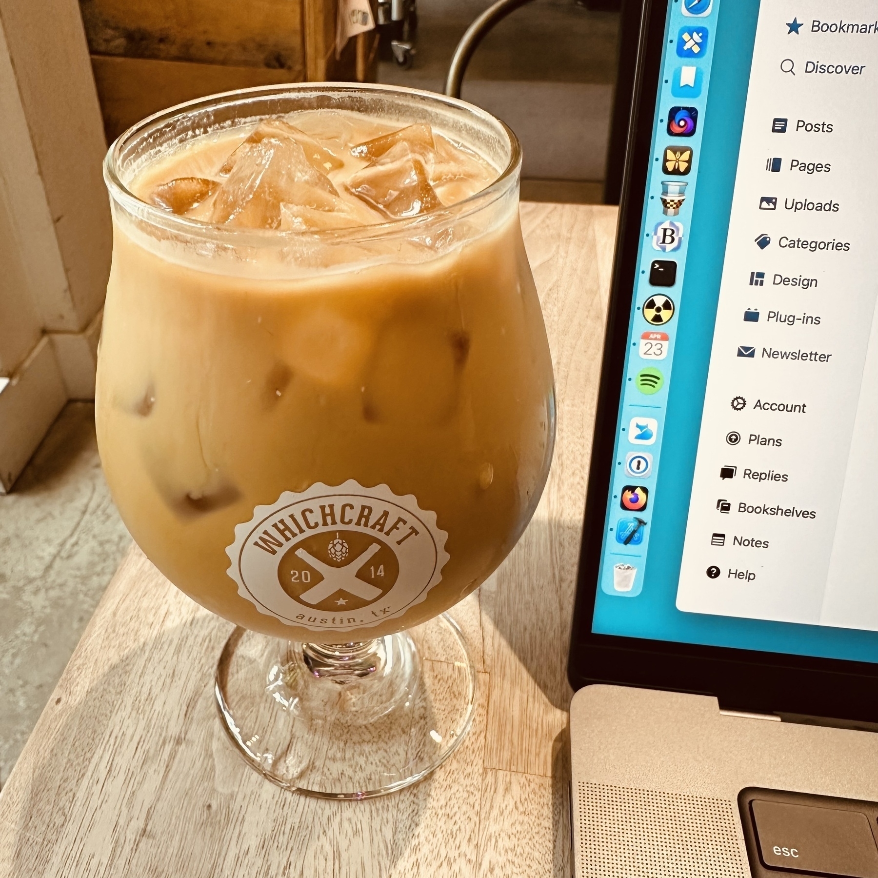 An iced latte in a nice glass next to a MacBook Pro with various Mac icons in the Dock, Micro.blog open in Safari, on a wooden table, with the word WhichCraft visible on the glass.