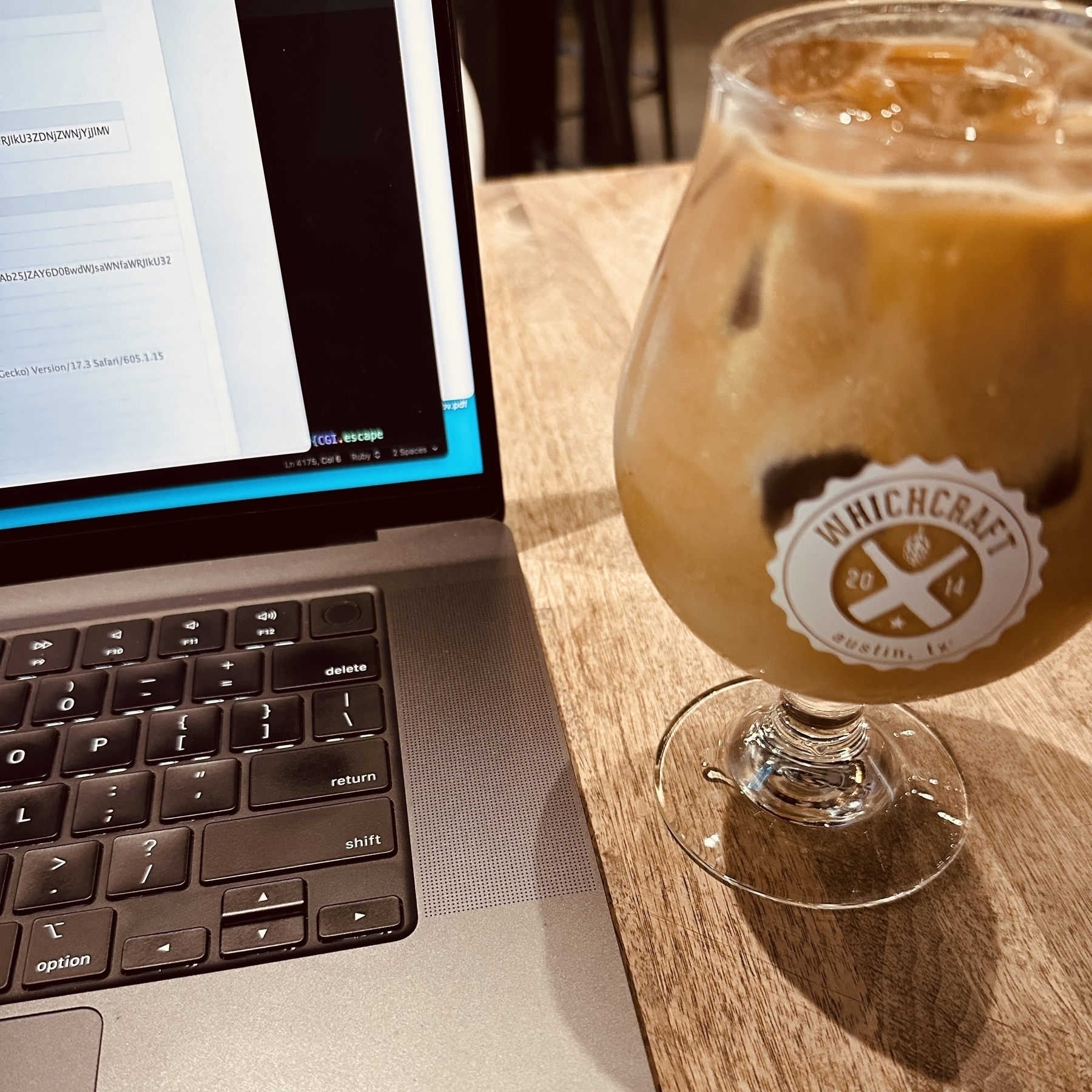 A glass of iced coffee sits on a table next to a laptop with text on its screen, part of a server message in Safari. The glass says Whichcraft.