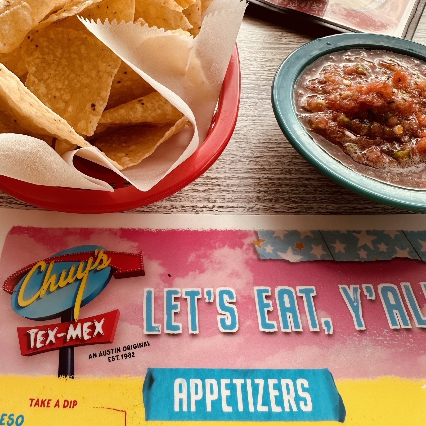 A bowl of salsa and a basket of tortilla chips, with the Chuy’s menu that says Let’s Eat, Y’all.