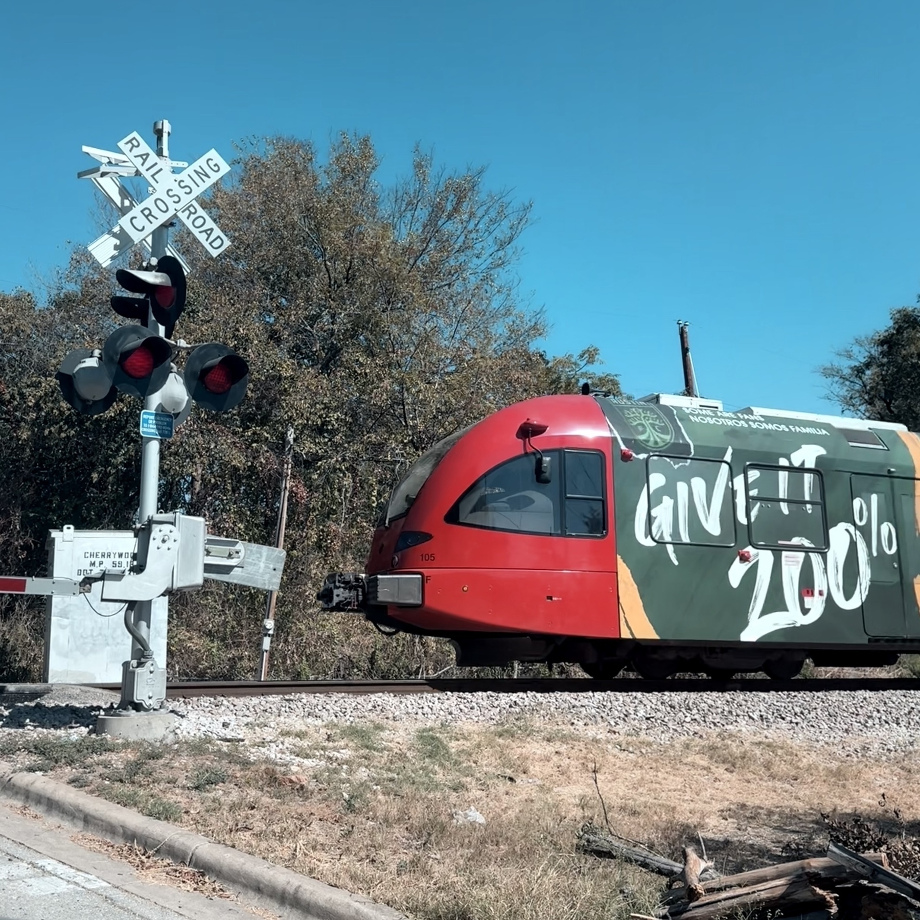 Austin MetroRail red train approaching railroad crossing with trees in background and blue sky.