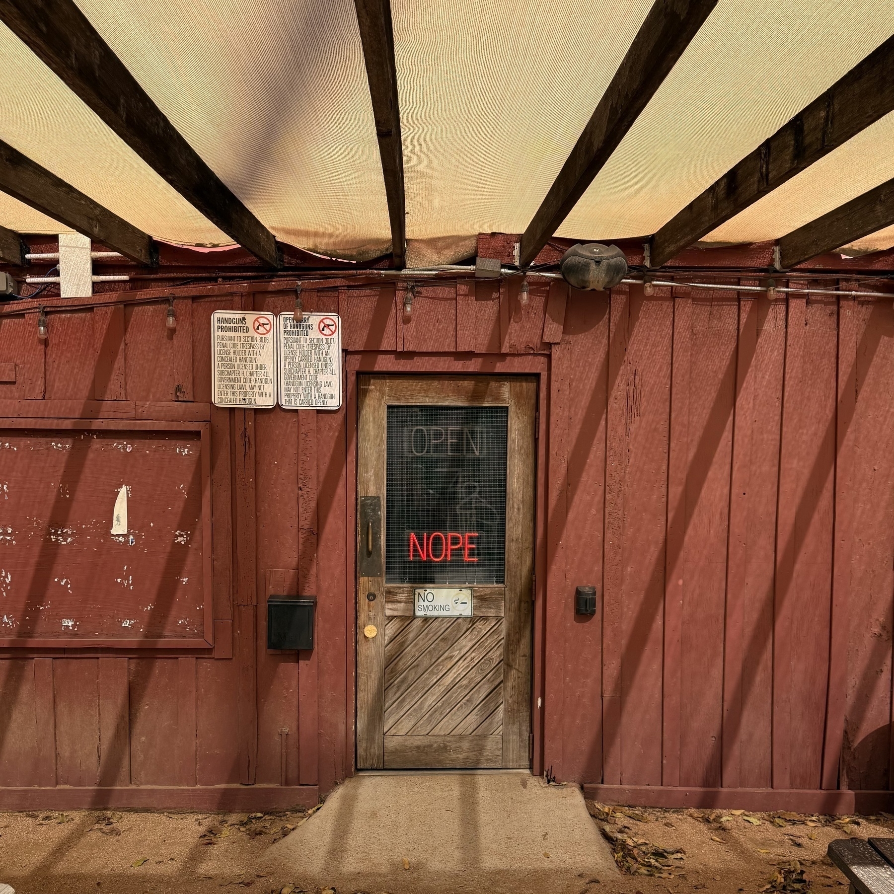 A rustic wooden door with a NOPE neon sign (OPEN is dimmed out) leads into a building shaded by a slatted canopy.