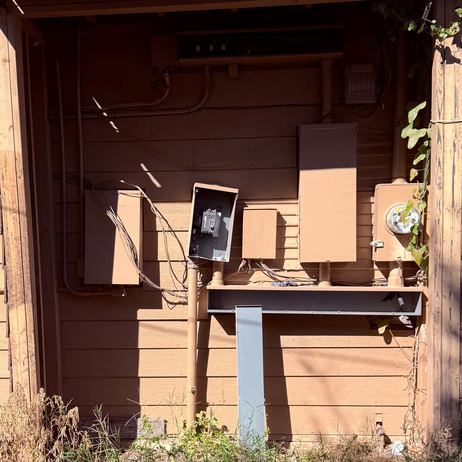 Electrical equipment and meters are mounted on a wooden wall, in disrepair, with surrounding grass and vines.