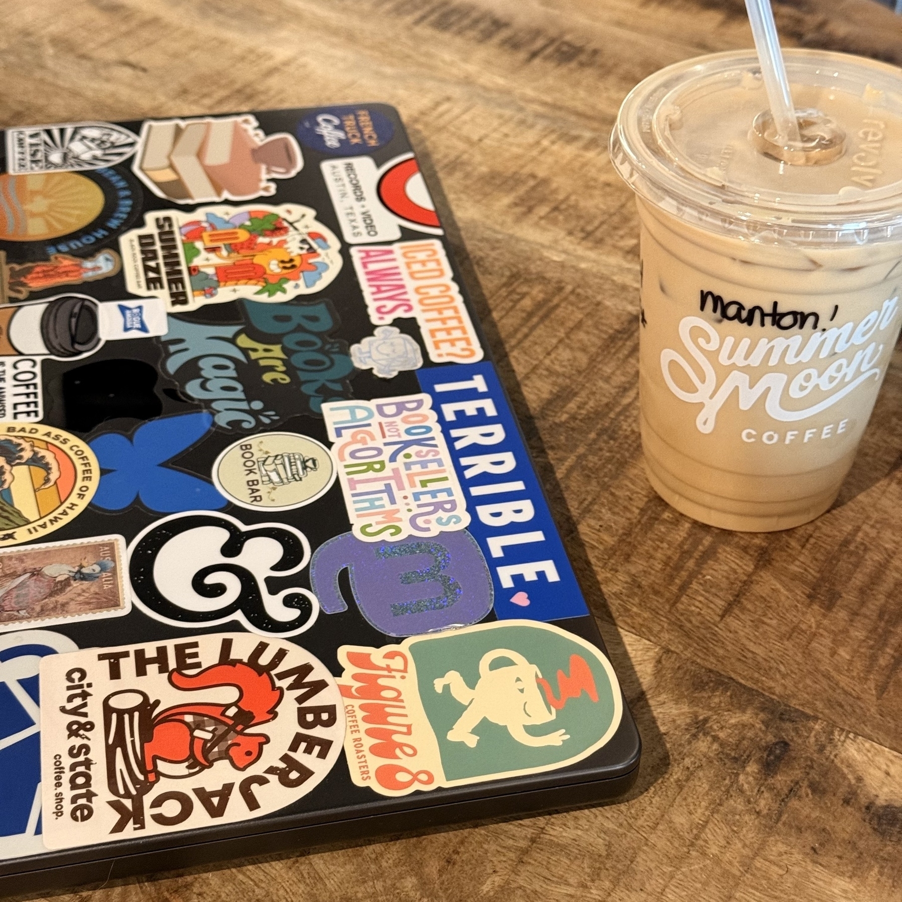 MacBook Pro covered in a variety of colorful stickers sits next to a plastic cup of iced coffee on a wooden table.