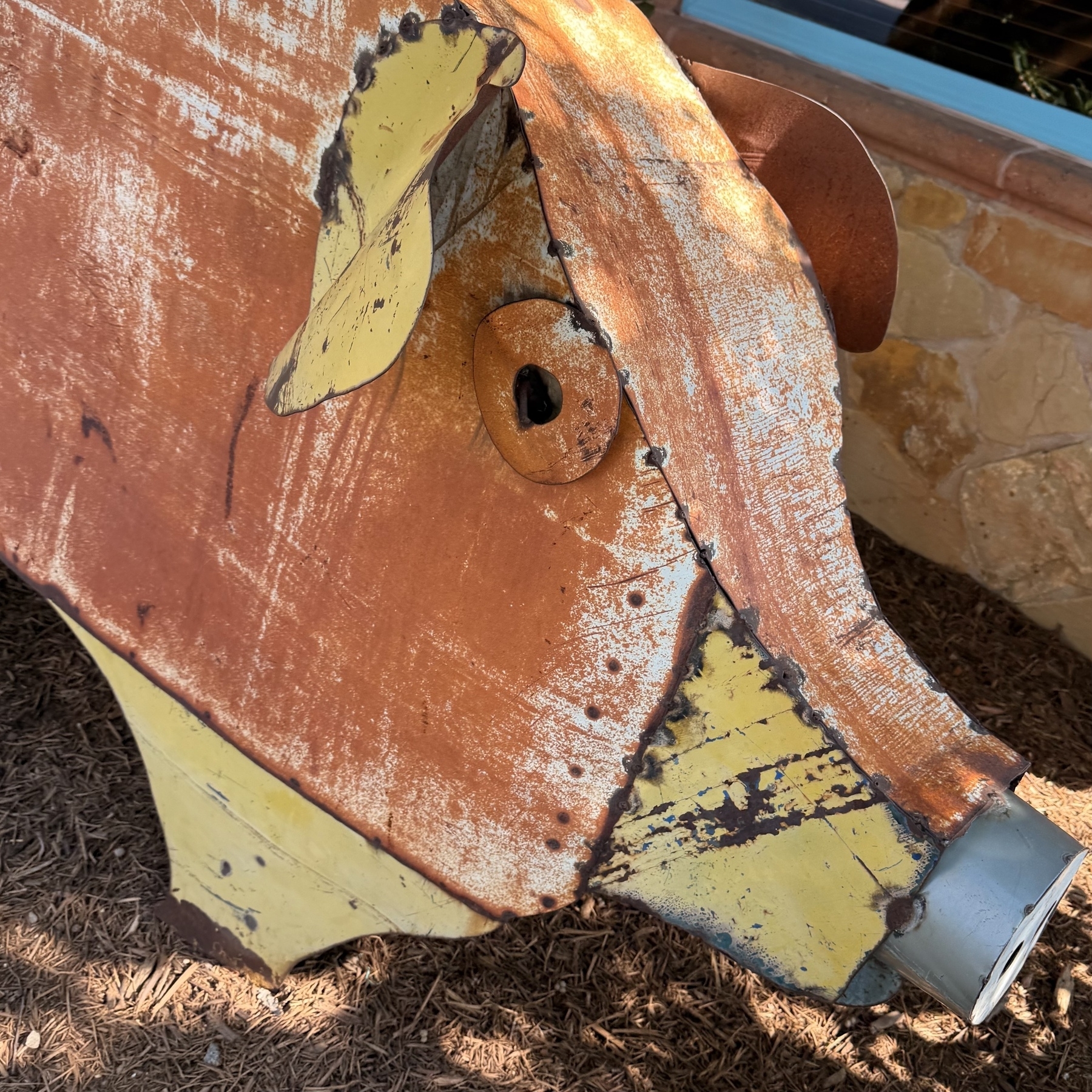 A large, rustic pig sculpture made of metal with a weathered orange and yellow finish stands on a ground covered in mulch.