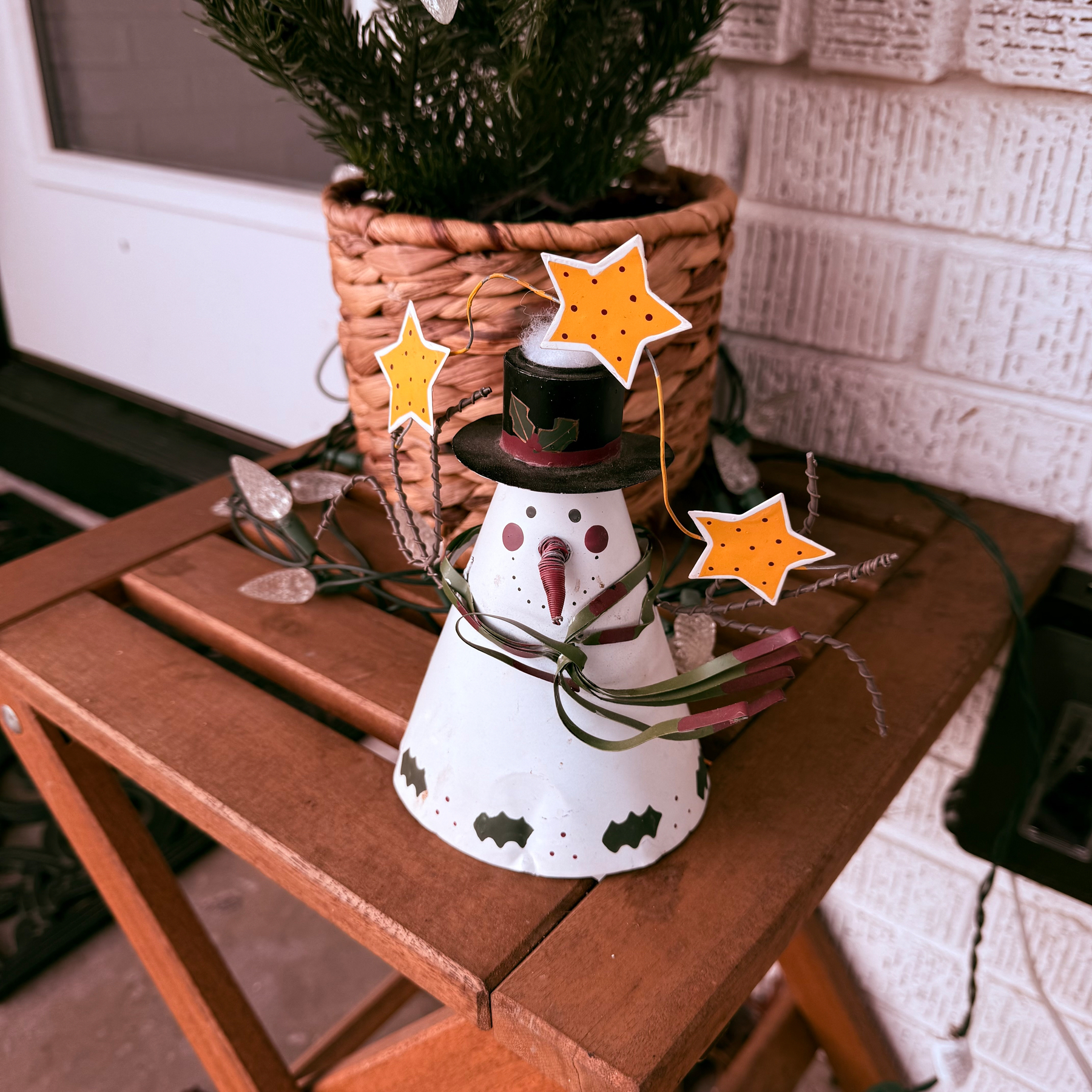 A small, festive snowman decoration with a top hat and orange stars is displayed on a wooden stool, next to a potted plant.