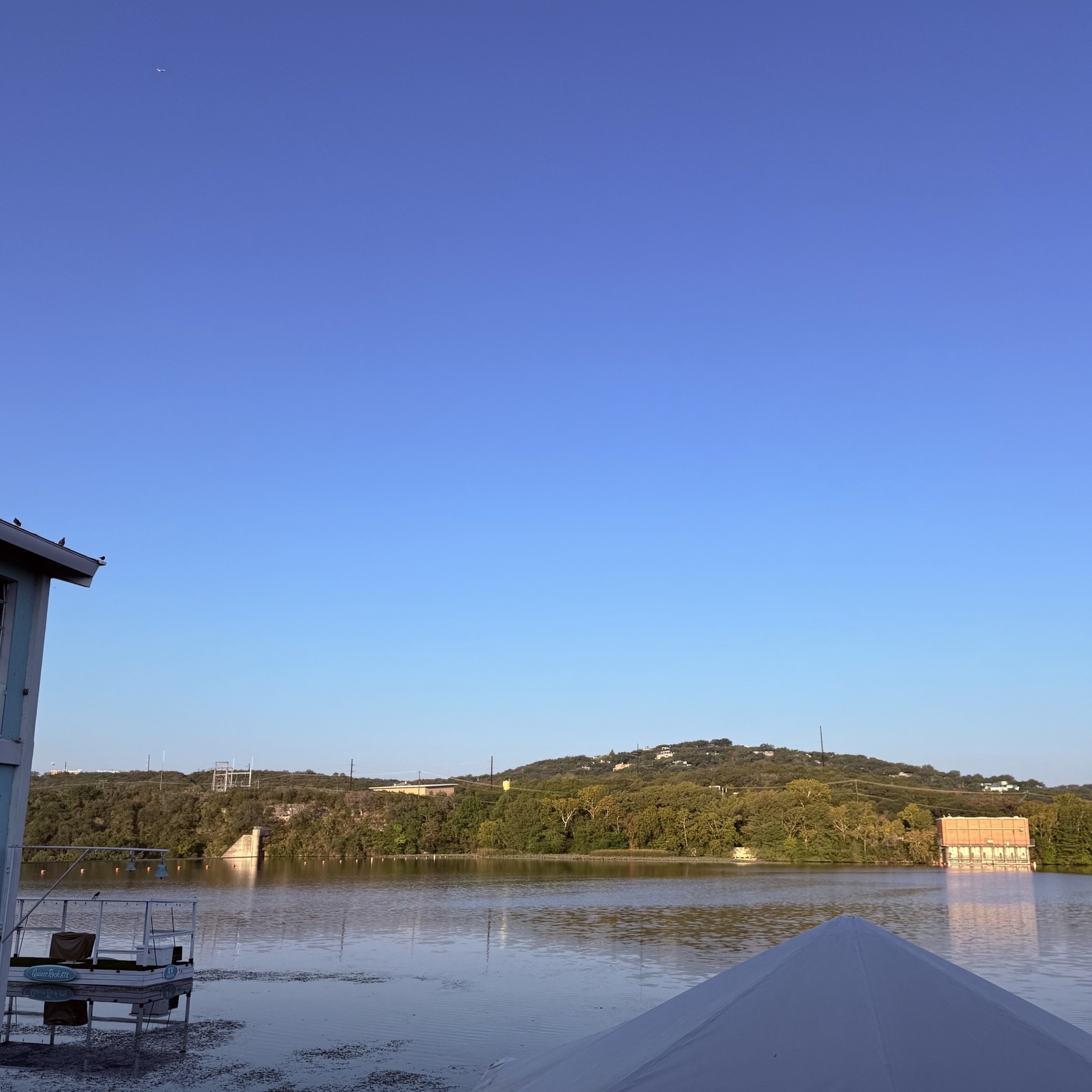 A calm river reflects surrounding trees and buildings under a clear blue sky.