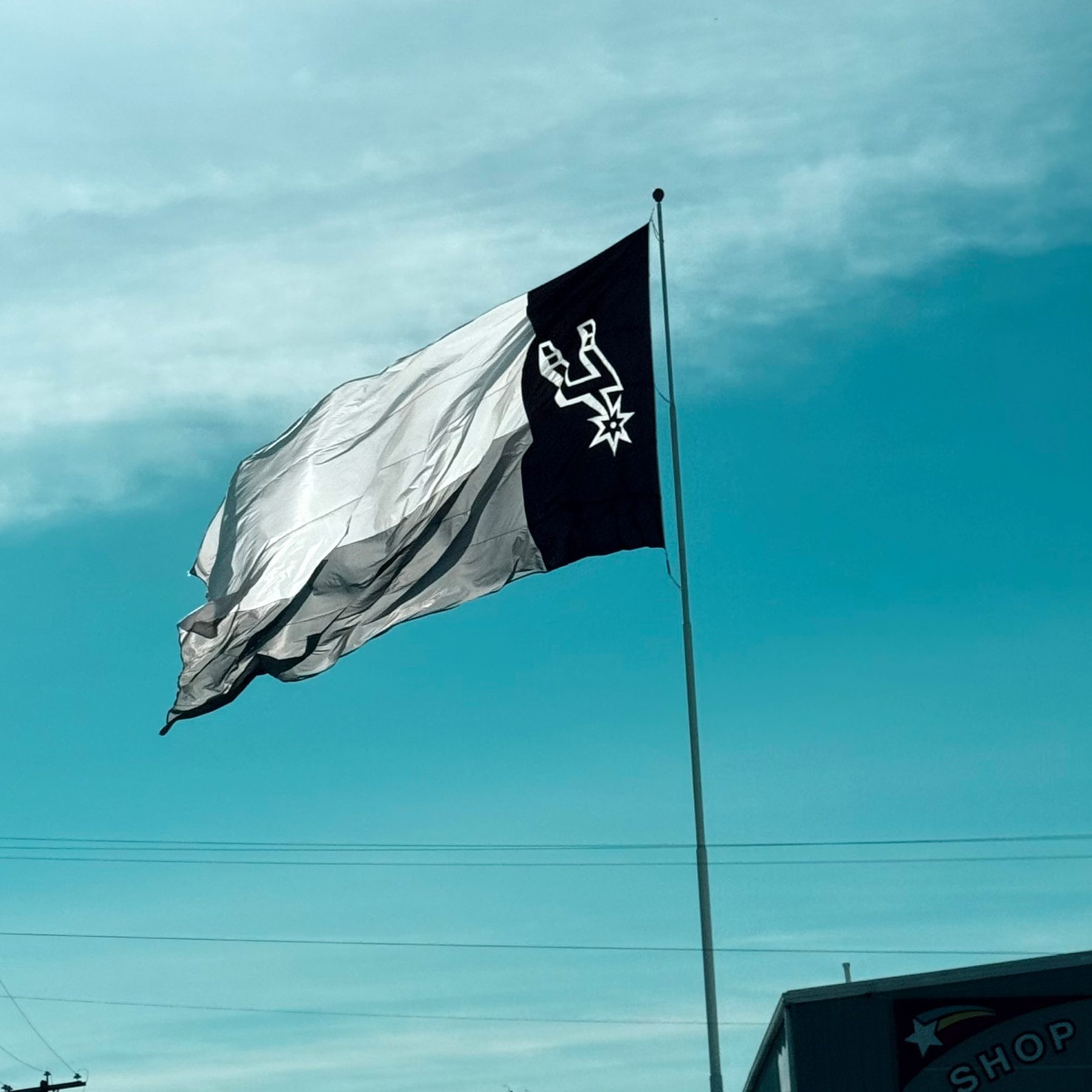 A black and white flag with the Spurs logo is waving against a blue sky.