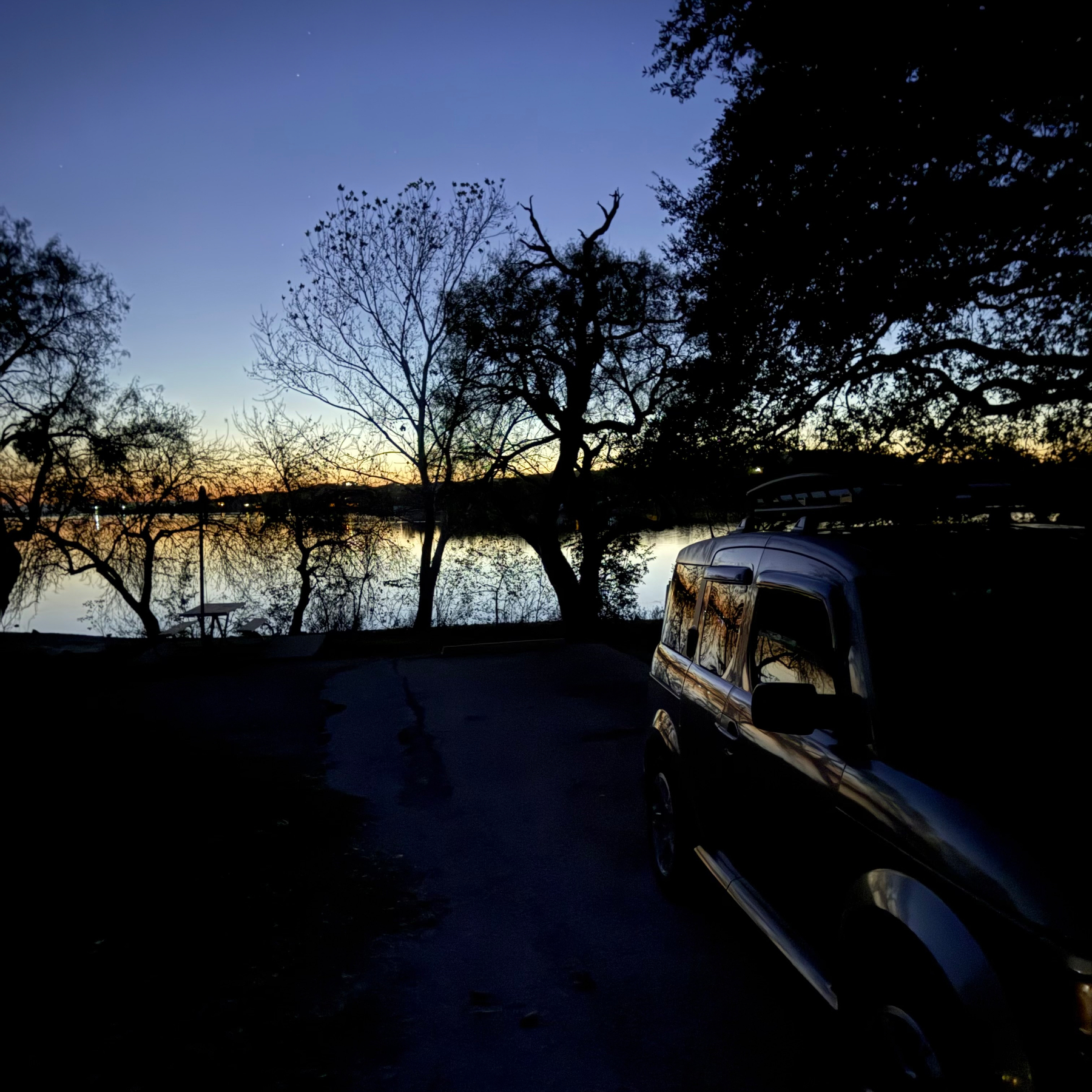 A car is parked near a serene lakeside at dusk, surrounded by silhouettes of trees against a dimly lit sky.