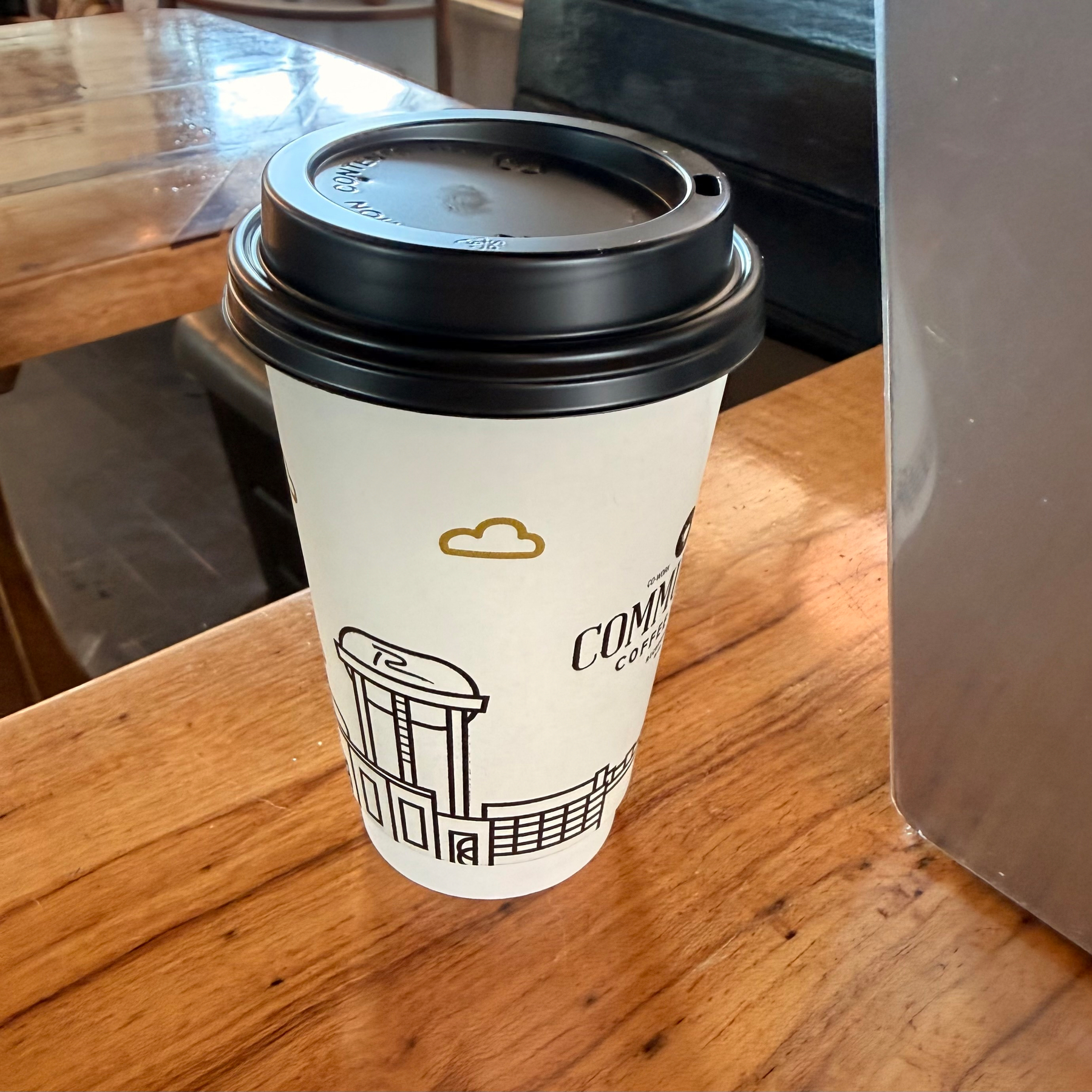 A coffee cup with a black lid and minimalist design sits on a wooden table in a cafe setting.