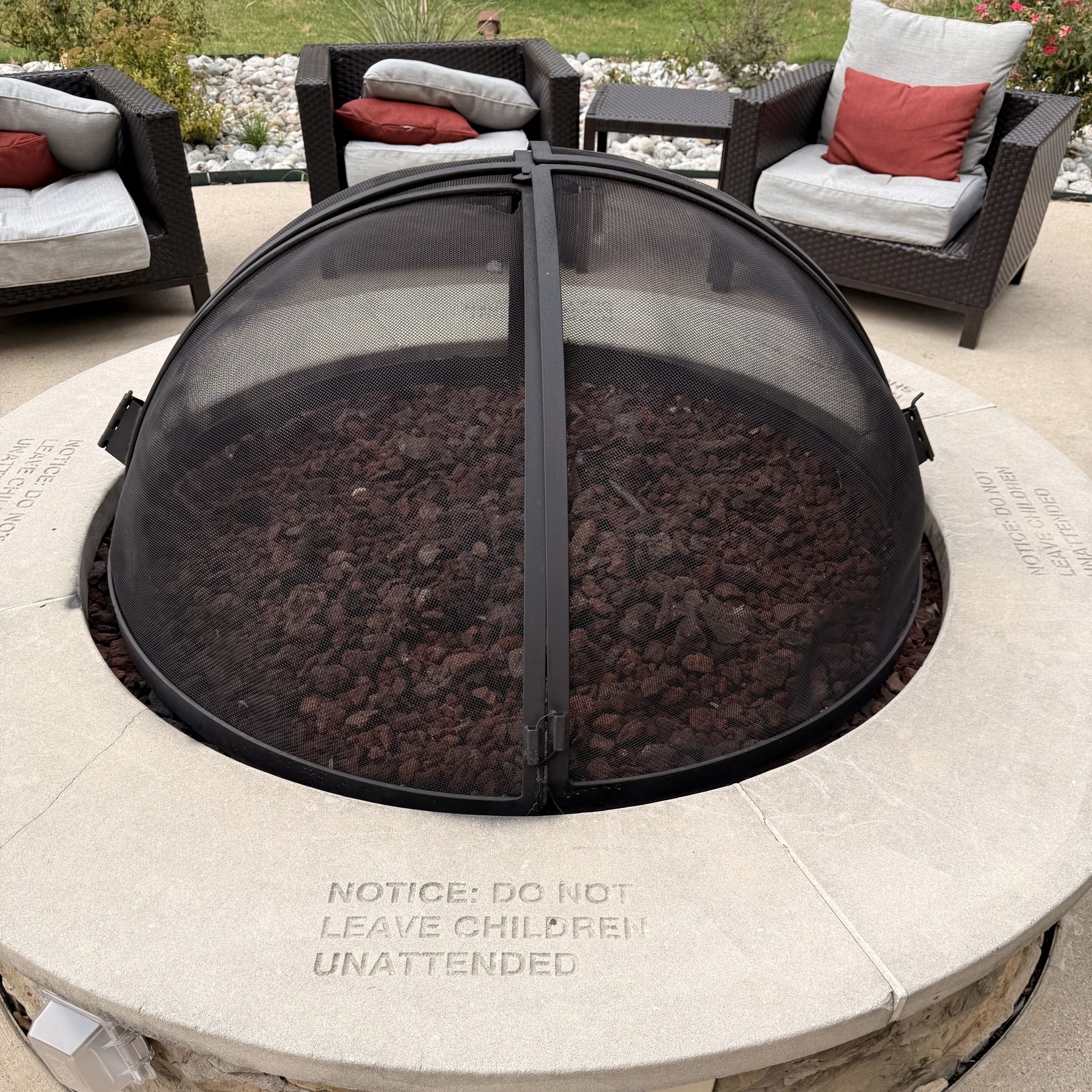 A covered fire pit is surrounded by cushioned outdoor chairs with a cautionary notice about watching children etched into the stone.