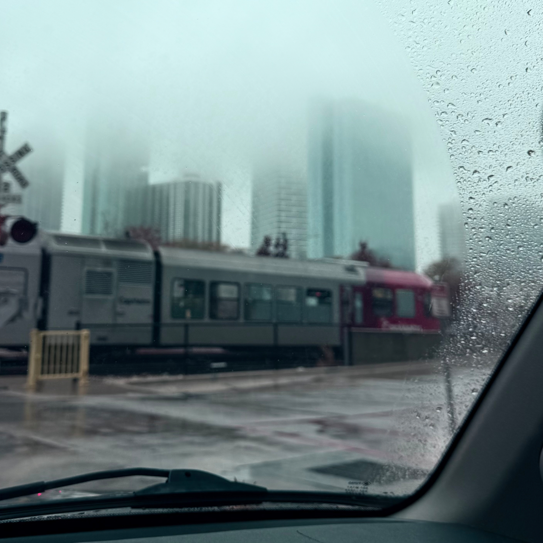 A train passes by a railroad crossing in a city with tall buildings shrouded in fog, viewed from inside a car.