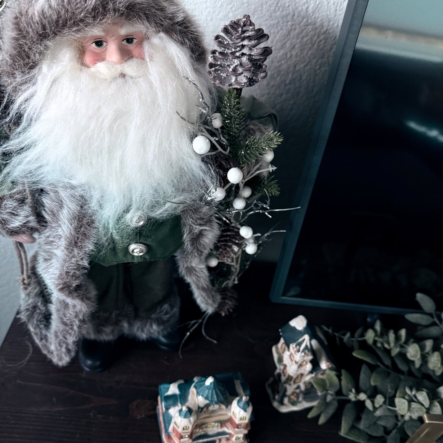 A Santa Claus figurine, dressed in a fur-trimmed outfit, holds a pinecone and greenery, standing next to a small model house and some decorative leaves.