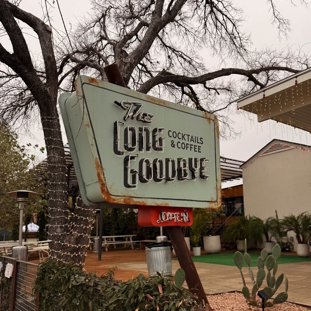 A vintage-looking sign for The Long Goodbye cocktail and coffee establishment stands outdoors surrounded by trees and desert plants.