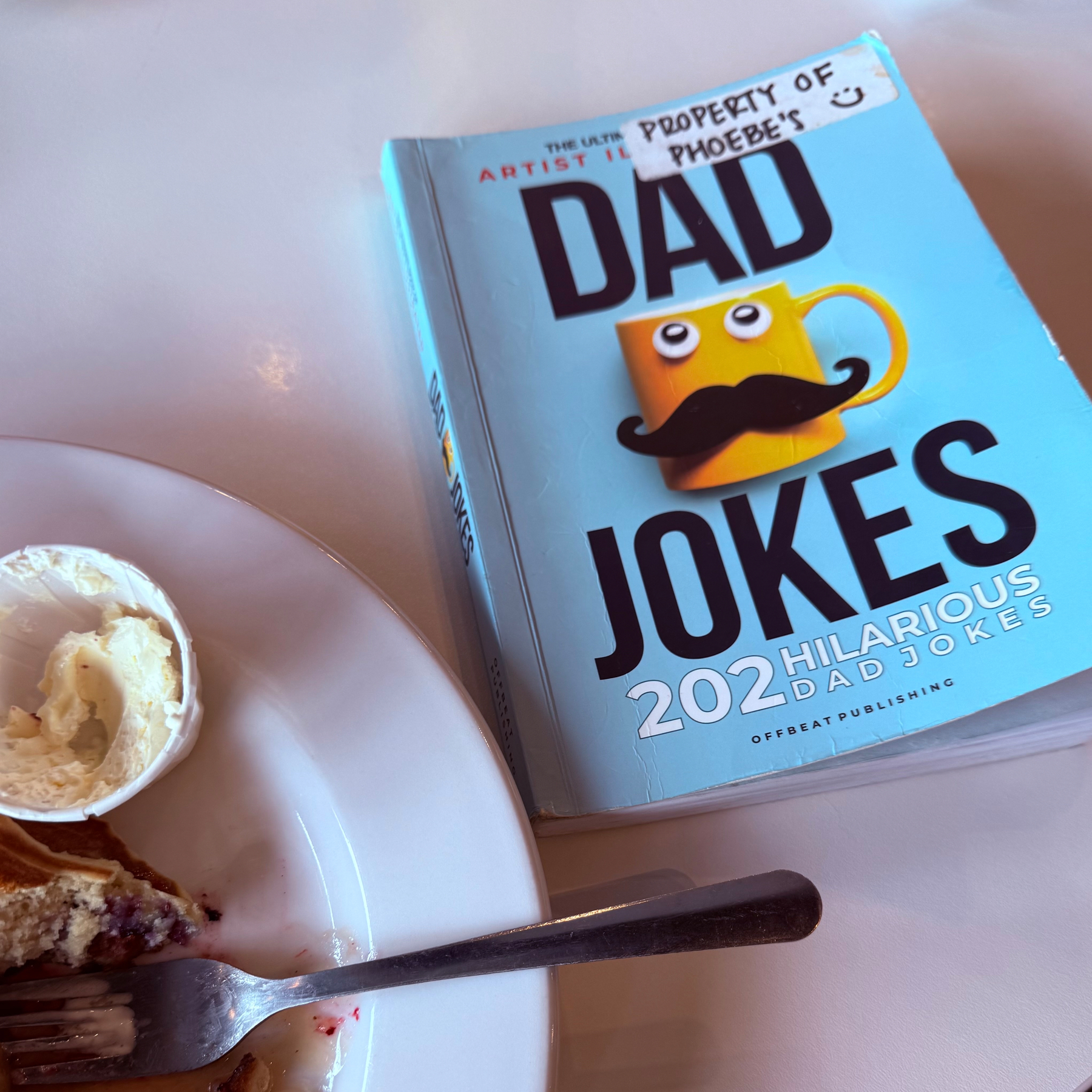 A book titled Dad Jokes: 202 Hilarious Dad Jokes sits on a table next to a plate with partially eaten pancakes.