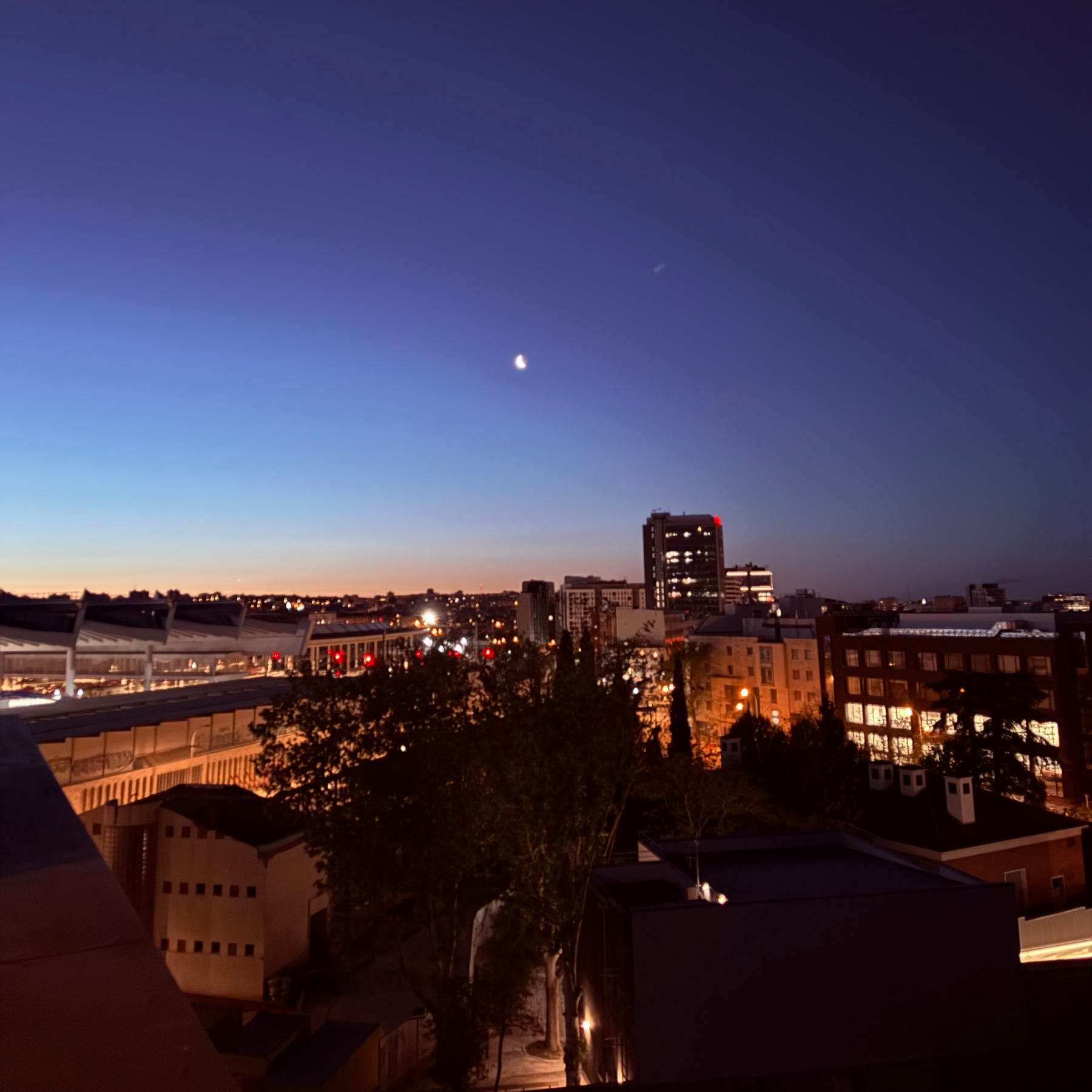 A cityscape at dusk features illuminated buildings, a crescent moon, and a dark sky.
