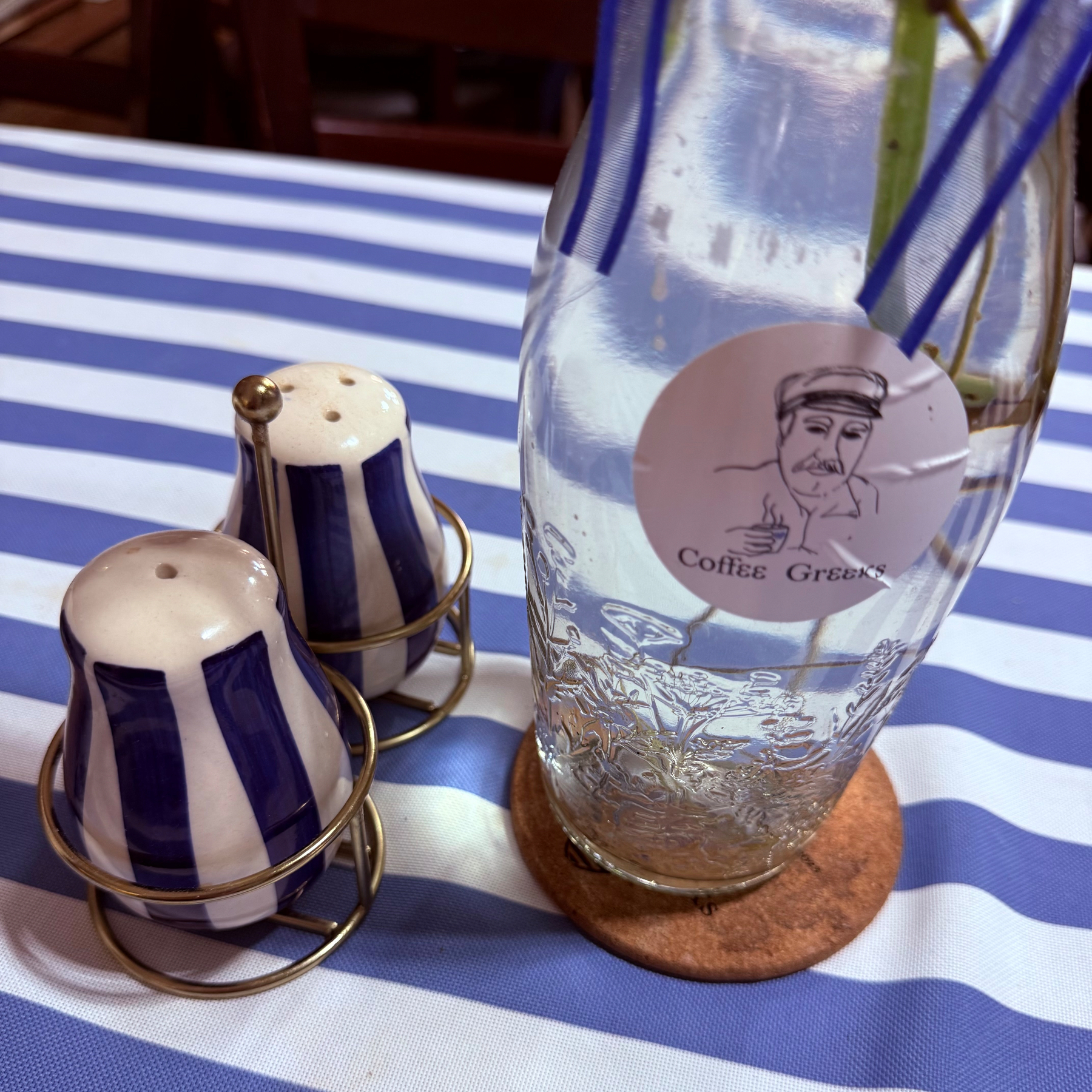 A blue-and-white striped salt and pepper shaker set sits next to a clear glass bottle with water and a Coffee Greeks label on a blue-and-white striped tablecloth.