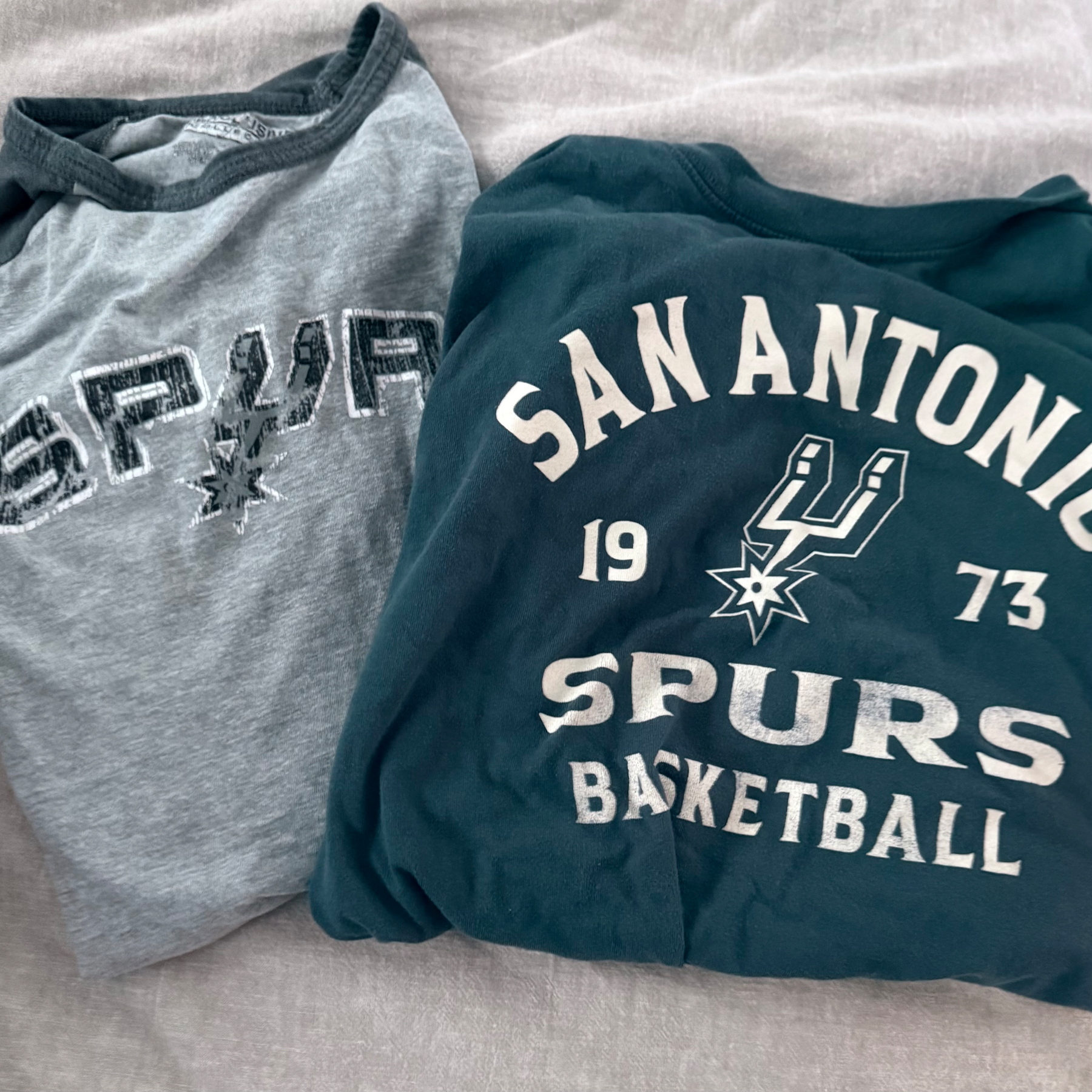 Two folded shirts featuring San Antonio Spurs logos and text are laid out on a surface.