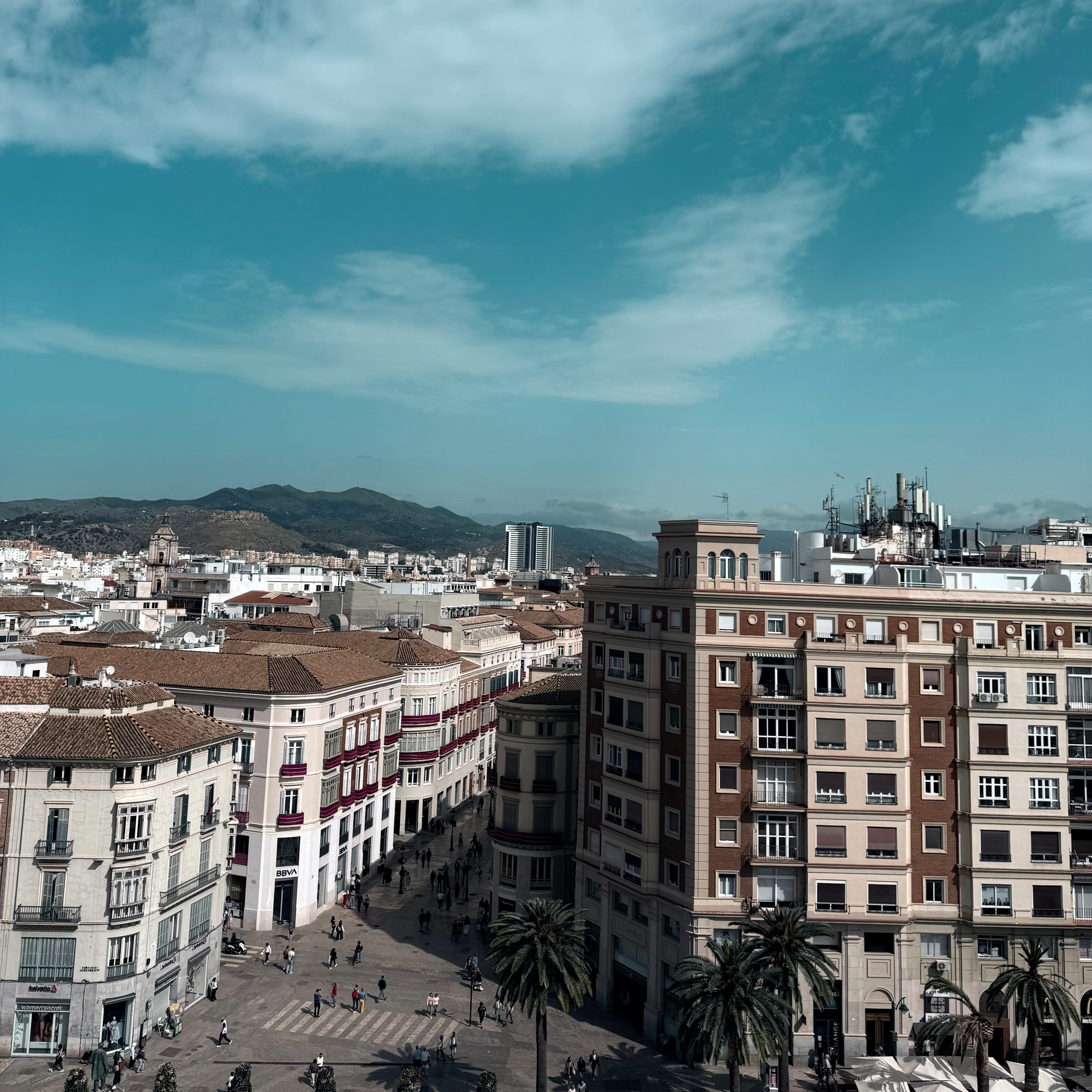 A scenic view of a city square surrounded by buildings under a partly cloudy sky, with mountains in the distance.