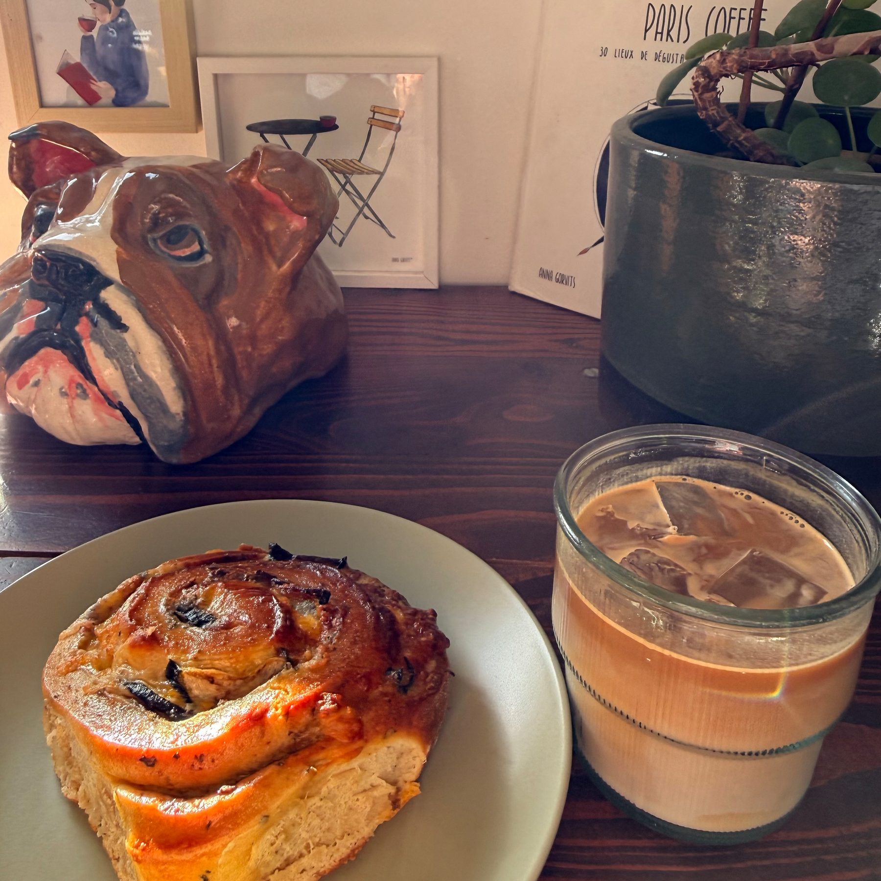 A ceramic bulldog, framed art, a potted plant, a pastry, and a glass of iced coffee are arranged on a wooden table.