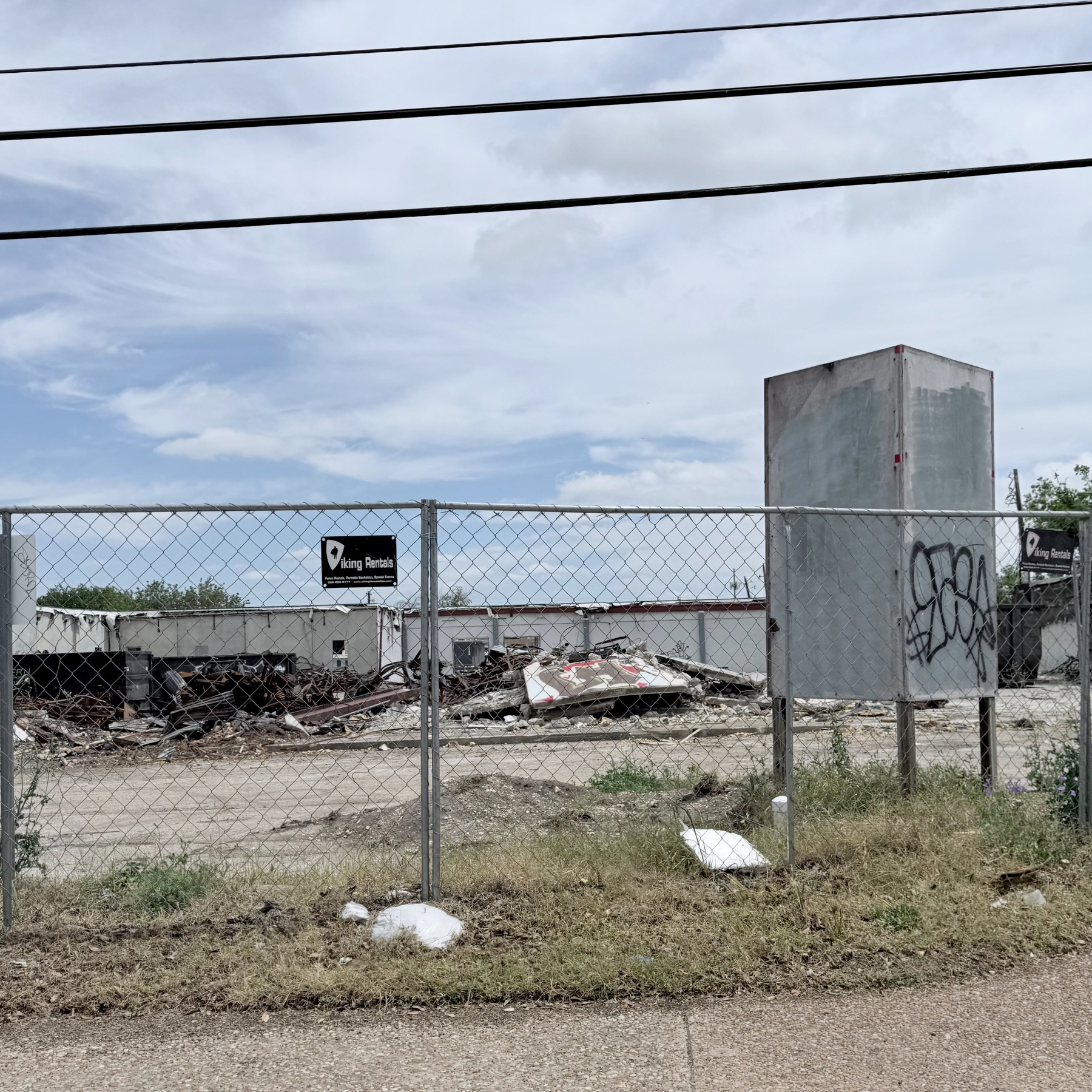 A chain-link fence stretches across the foreground under a cloudy sky, with an old building and rubble in the background.