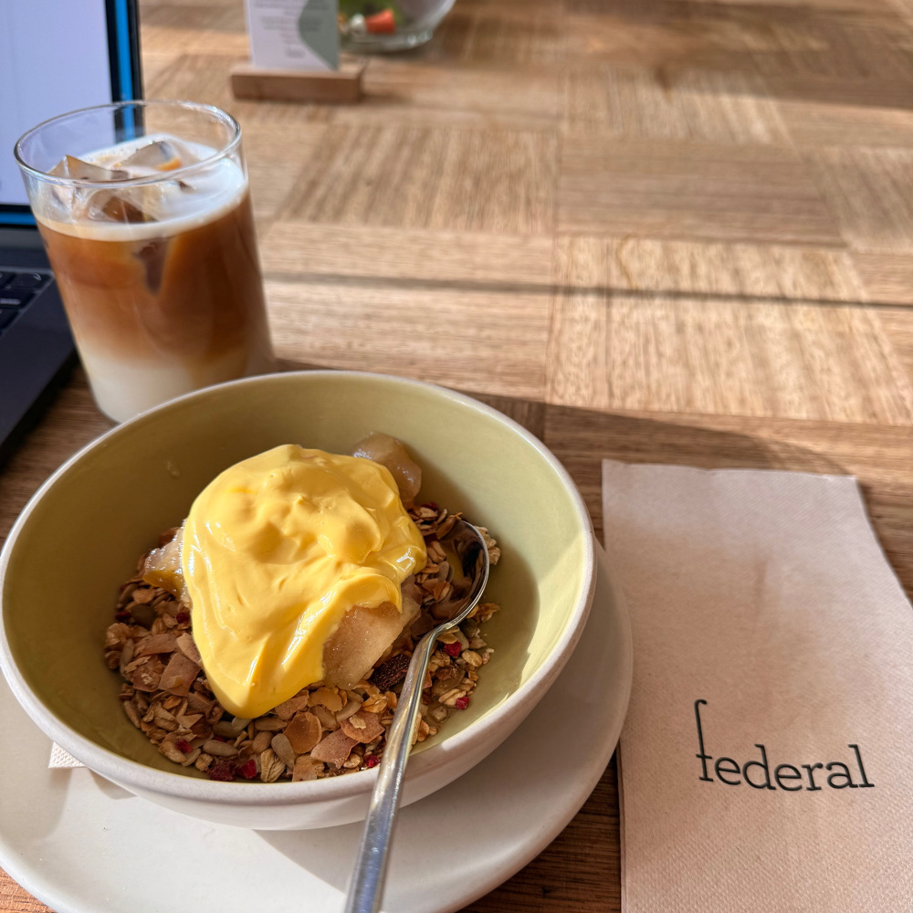 A bowl of muesli topped with a yogurt and served with iced coffee sits on a table next to a napkin labeled federal.