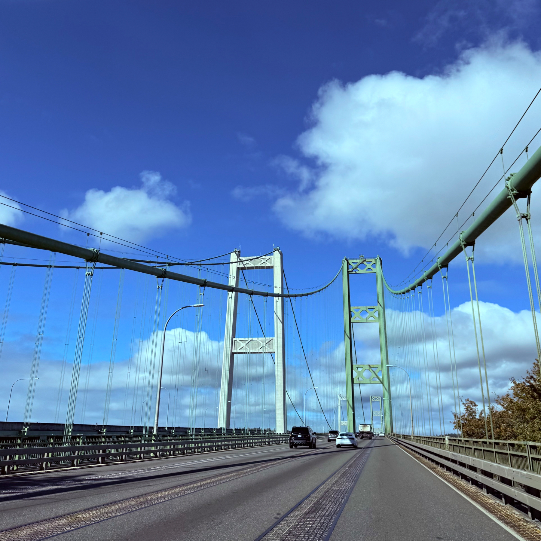 A suspension bridge stretches across a clear blue sky with a few white clouds, supporting vehicles traveling along the roadway.