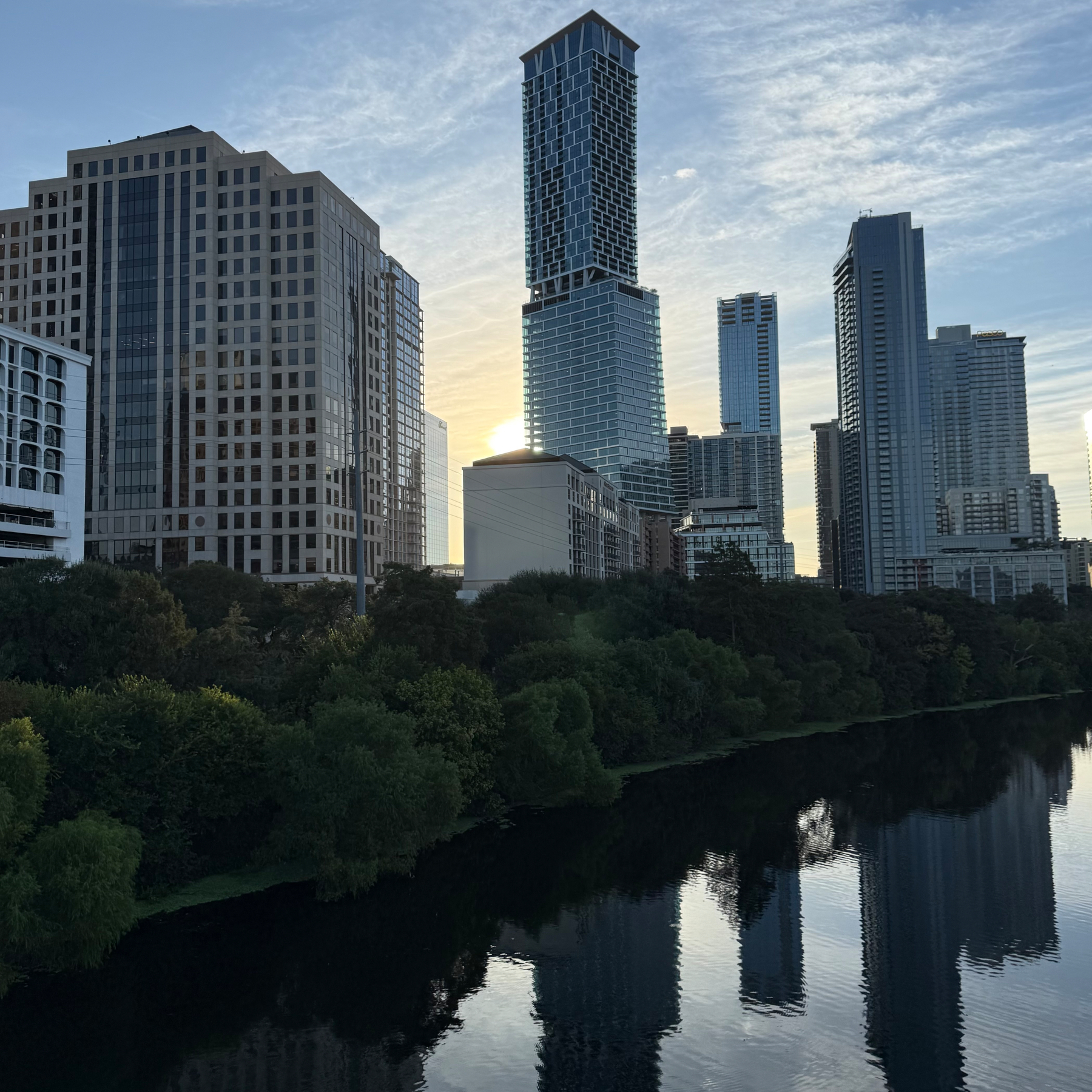 A city skyline with tall buildings is reflected in a calm body of water, with trees lining the waterfront under a partly cloudy sky.