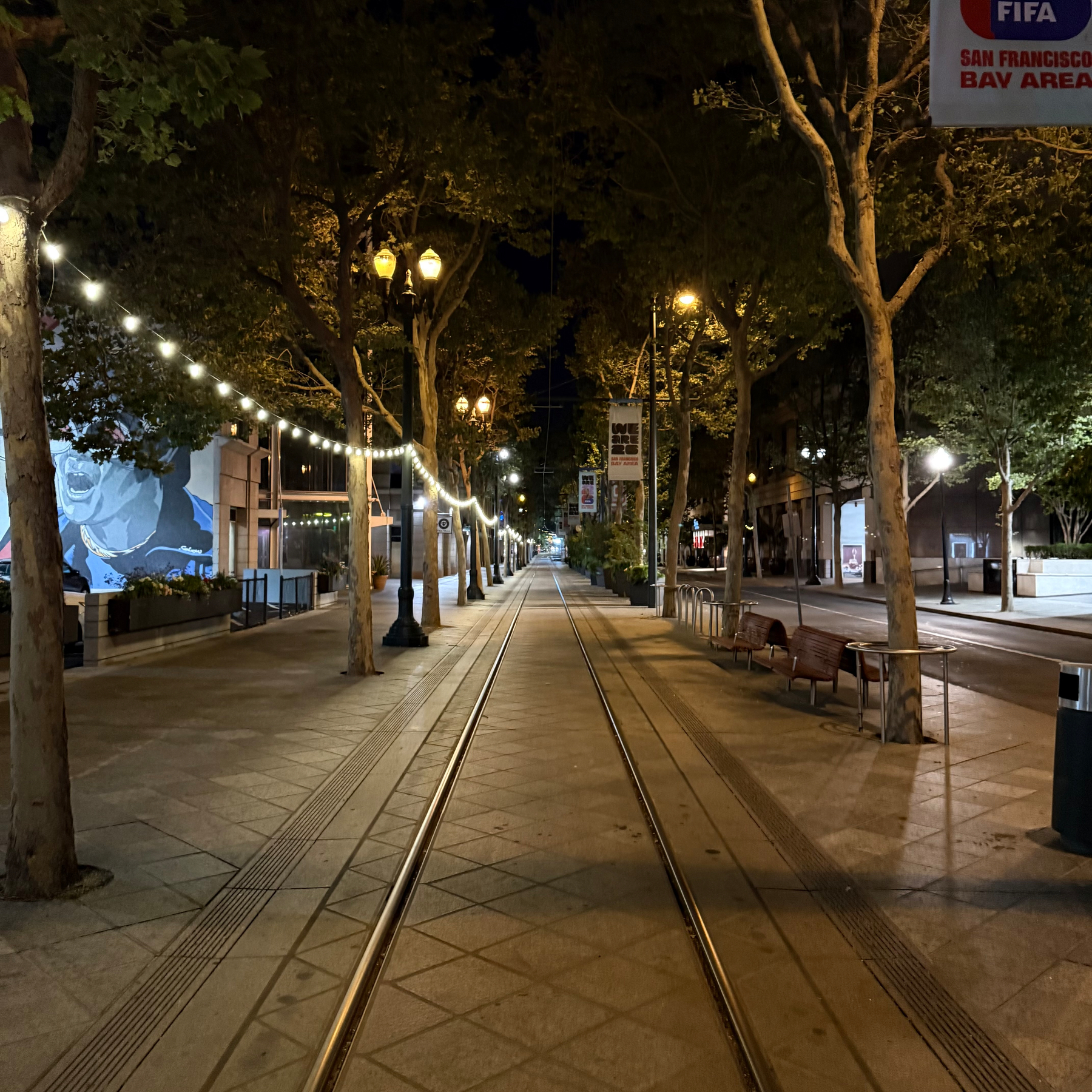 A well-lit, empty street at night with tram tracks running down the center, lined with trees and benches.