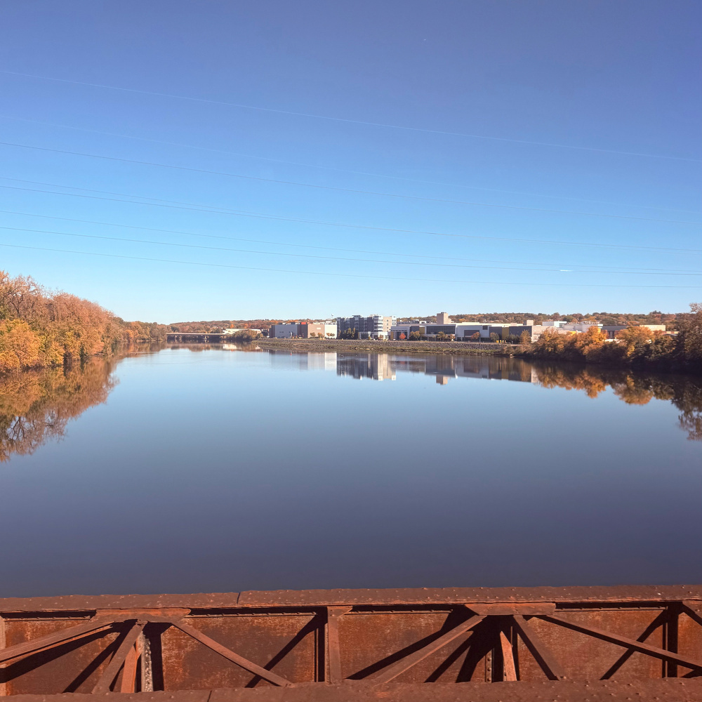 A calm river reflects the clear blue sky, autumn-colored trees, and distant buildings.