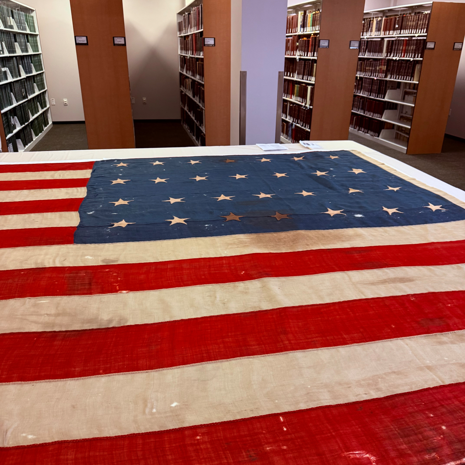 An American flag is displayed flat on a table in a library surrounded by shelves filled with books.