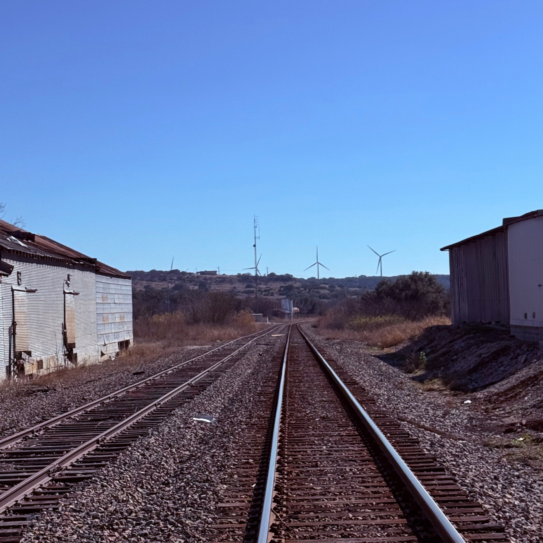 Railroad tracks stretch into the distance flanked by industrial buildings, with wind turbines visible on the horizon.