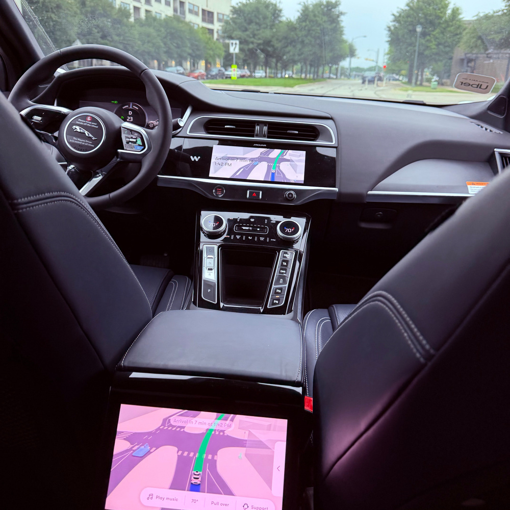 The photo shows the interior of a Jaguar car, featuring a steering wheel with the logo, multiple screens displaying navigation maps, and a view through the windshield of a road lined with trees. No one is driving the car.