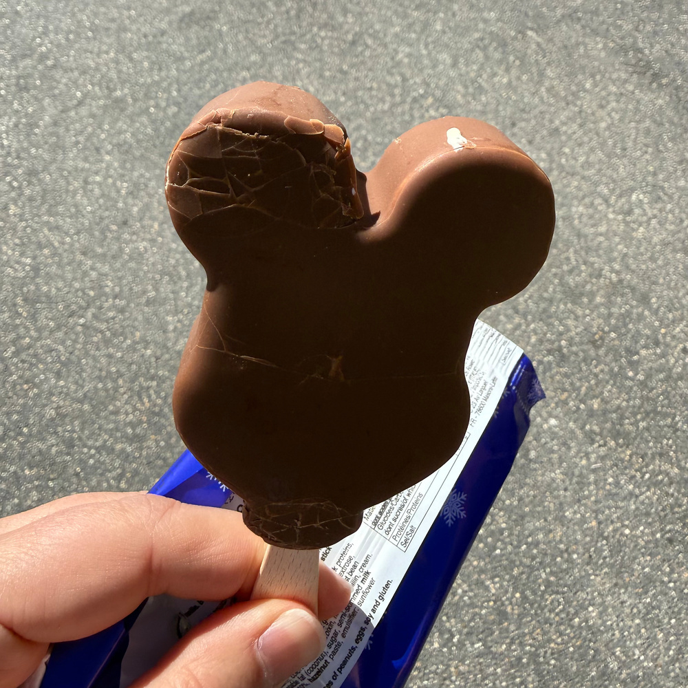 A hand is holding a chocolate ice cream bar shaped like a cartoon character's head, partially unwrapped.