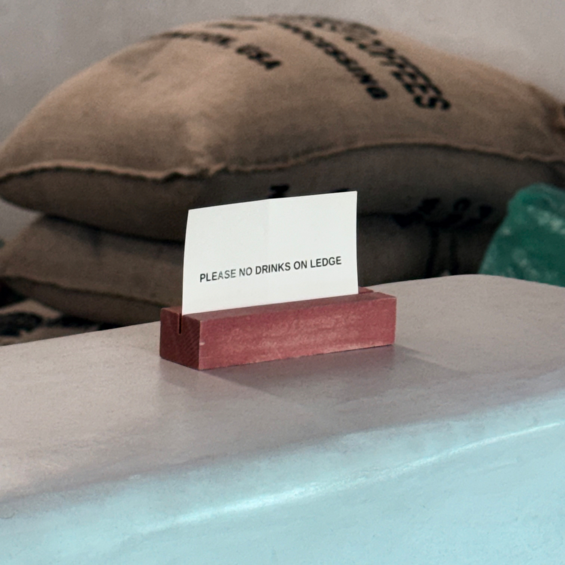 A sign on a ledge reads Please No Drinks On Ledge, with sacks and a green bag in the background.