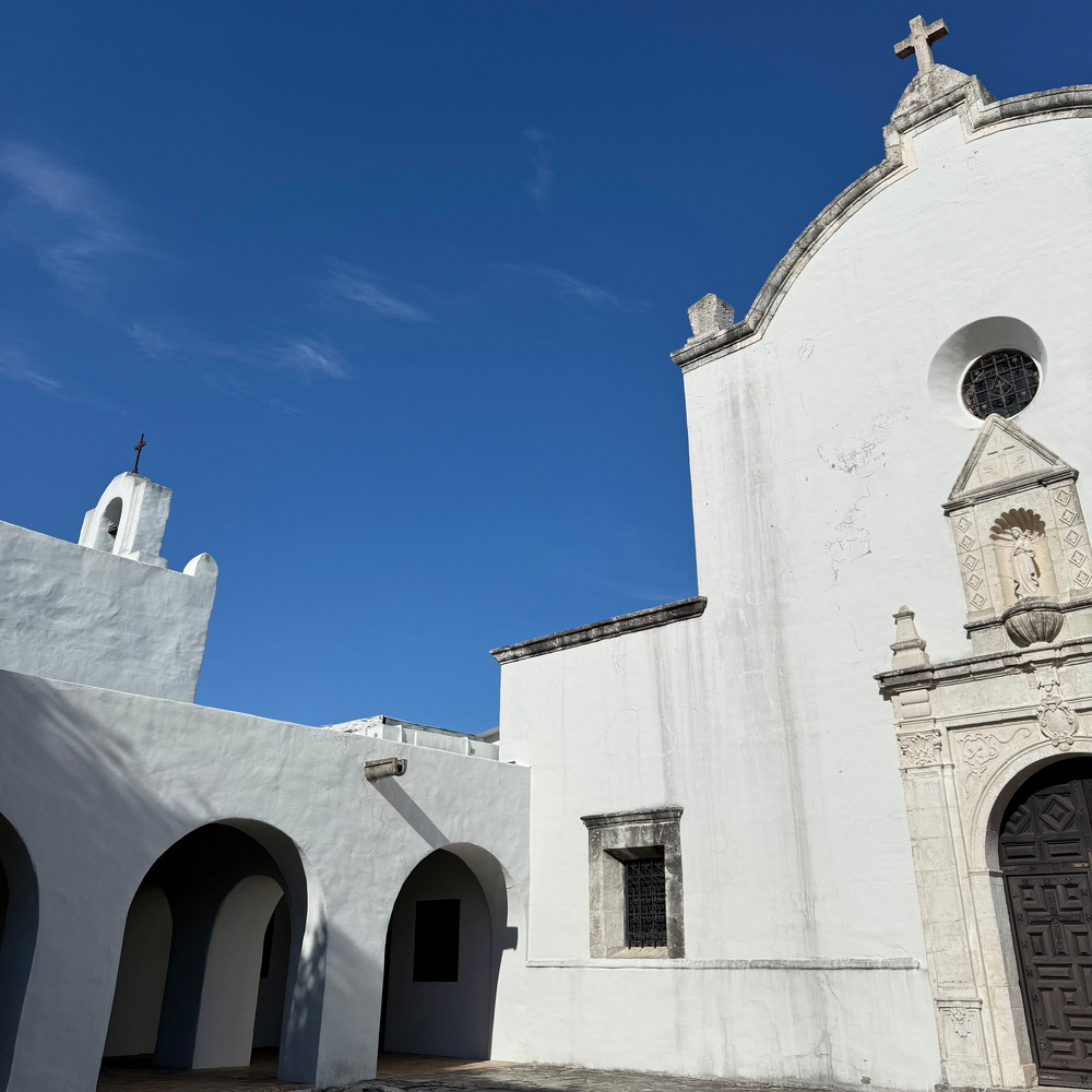 Auto-generated description: A white colonial-style building with arches, a cross on top, and ornate entrance details stands against a clear blue sky.
