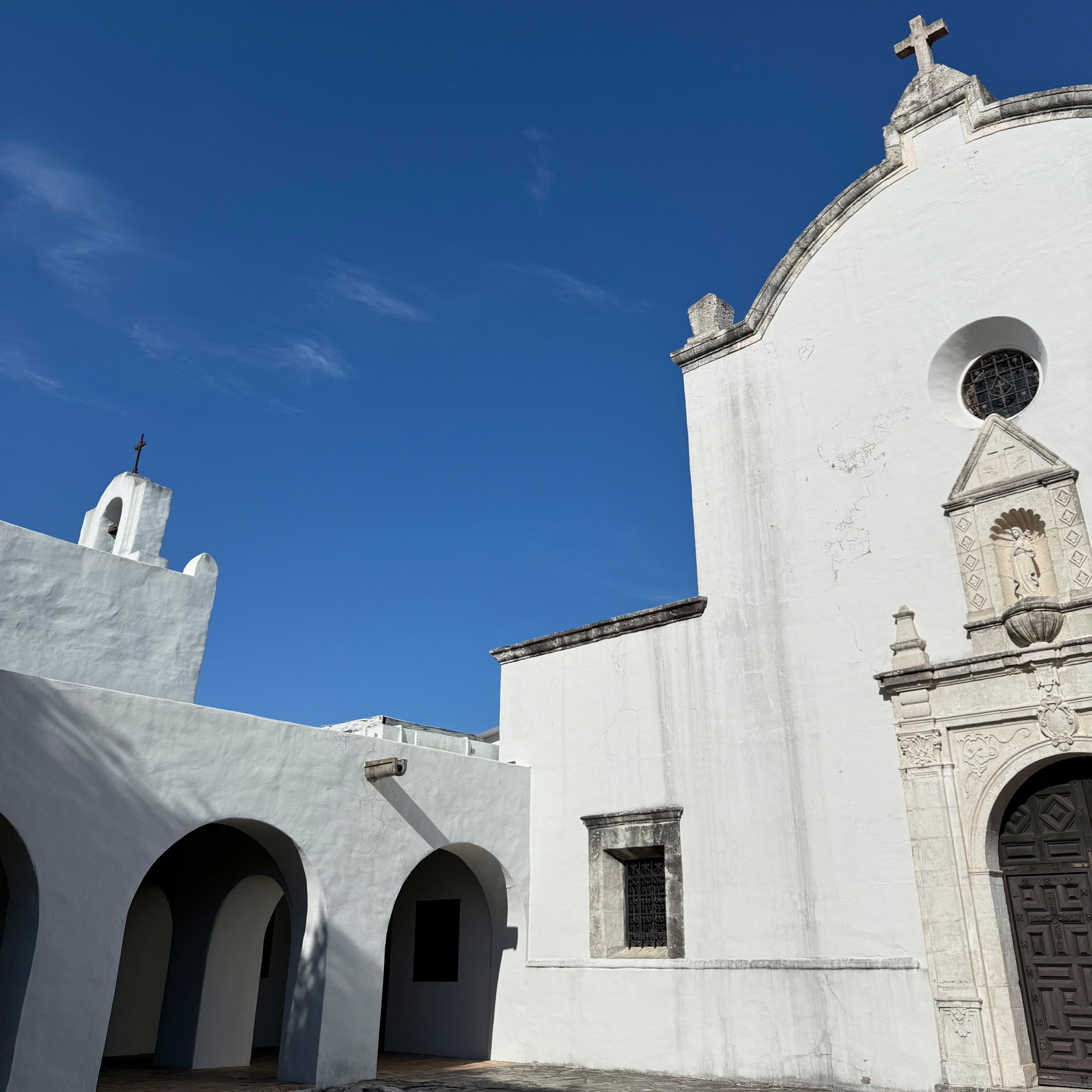 Auto-generated description: A white colonial-style building with arches, a cross on top, and ornate entrance details stands against a clear blue sky.