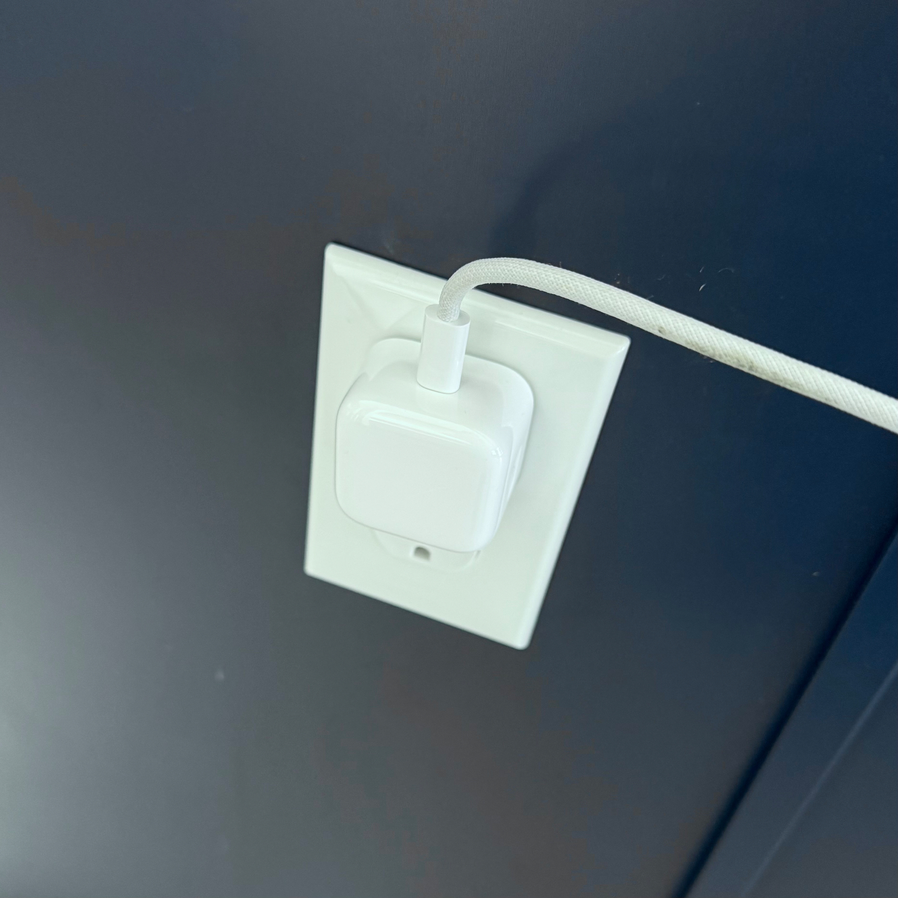 A white plug with a cable is inserted into a wall socket on a dark blue wall.