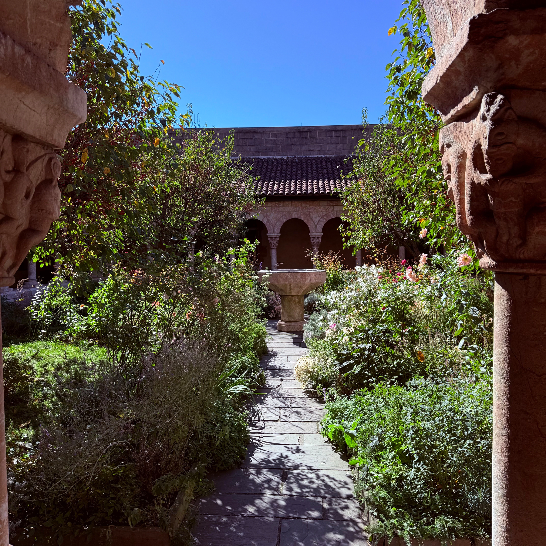 A stone pathway leads through a lush garden courtyard with various plants and flowers, framed by ancient stone columns and arches under a clear blue sky.