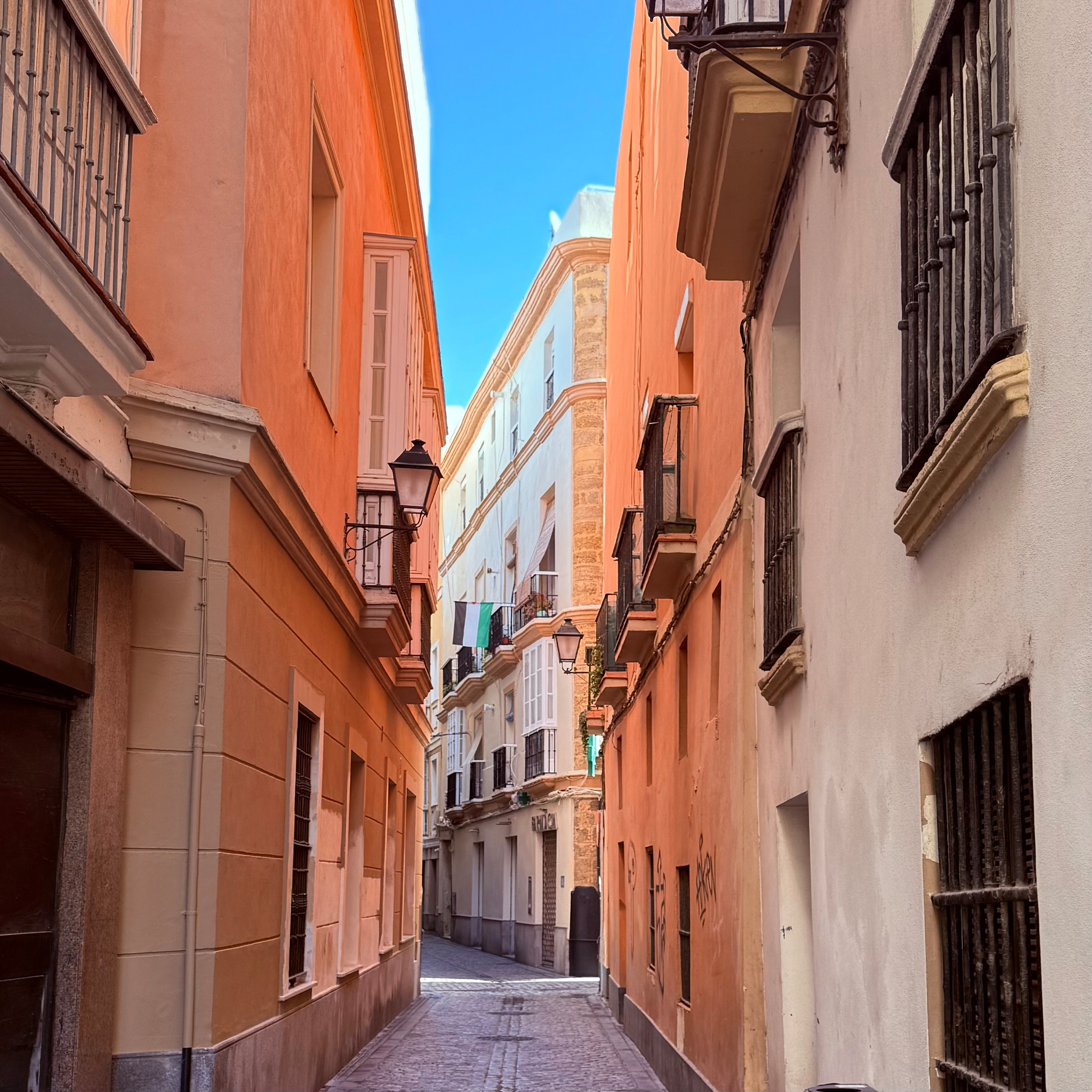 A narrow, cobblestone alley is bordered by colorful, traditional European buildings with balconies on either side, leading into the distance under a clear blue sky.