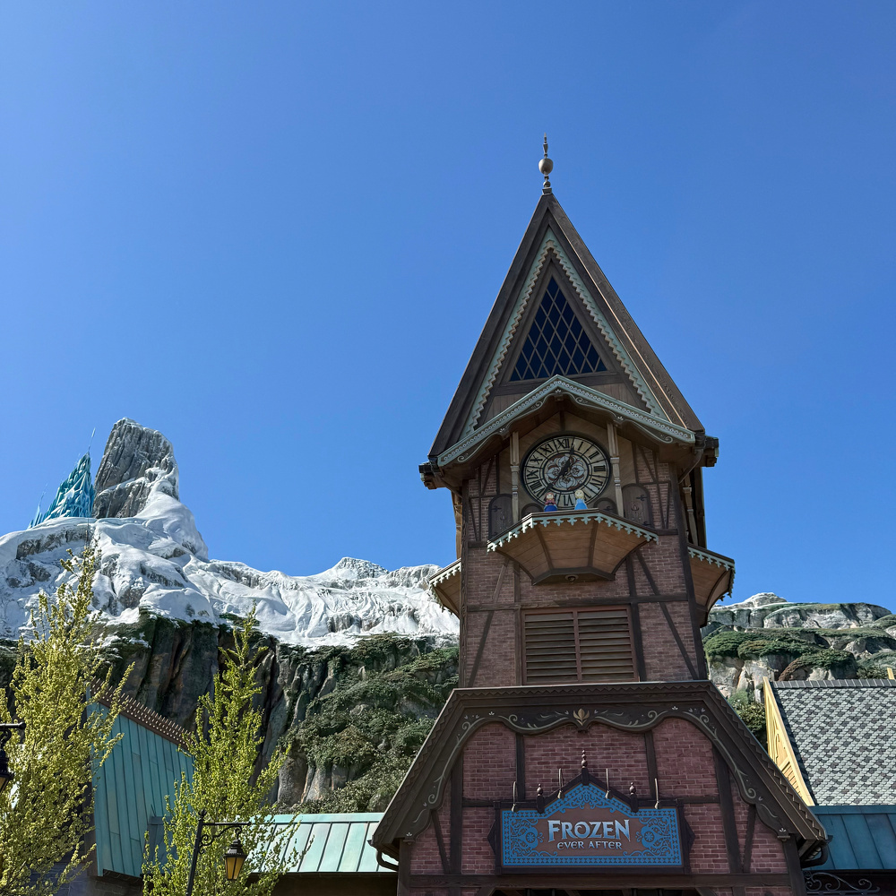 A quaint clock tower stands against the backdrop of snow-covered mountains under a clear blue sky.