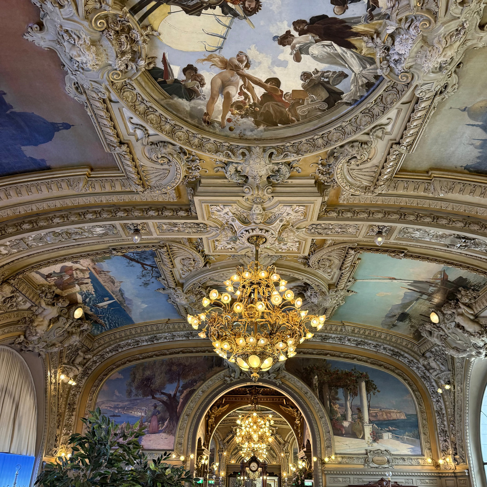 A luxurious room features ornate ceilings with detailed paintings and an elaborate chandelier.