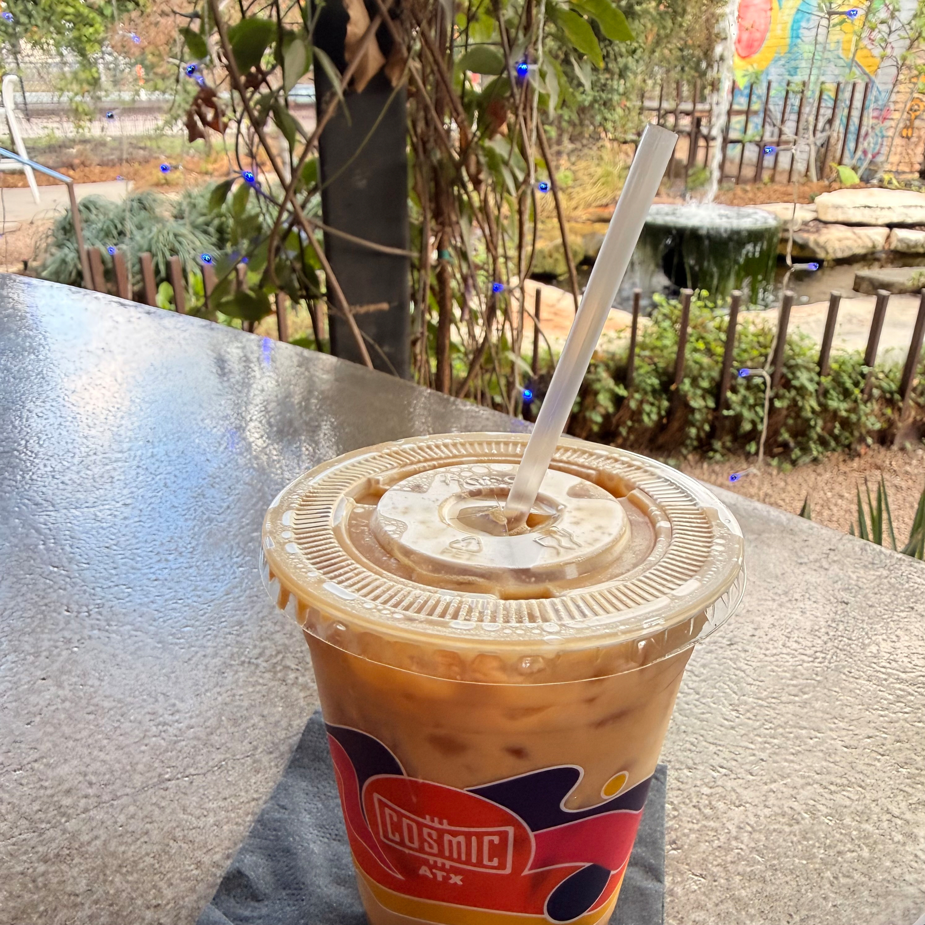A plastic cup with iced coffee sits on a table near a garden setting with a pond in the background.