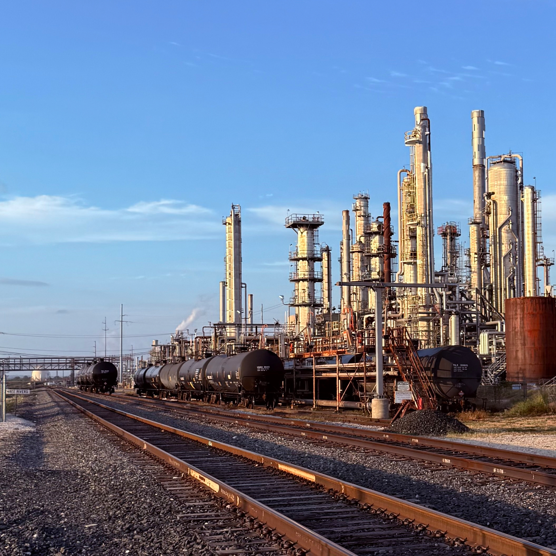 Railroad tracks run alongside an industrial refinery complex with tall distillation towers and several black tanker railcars.