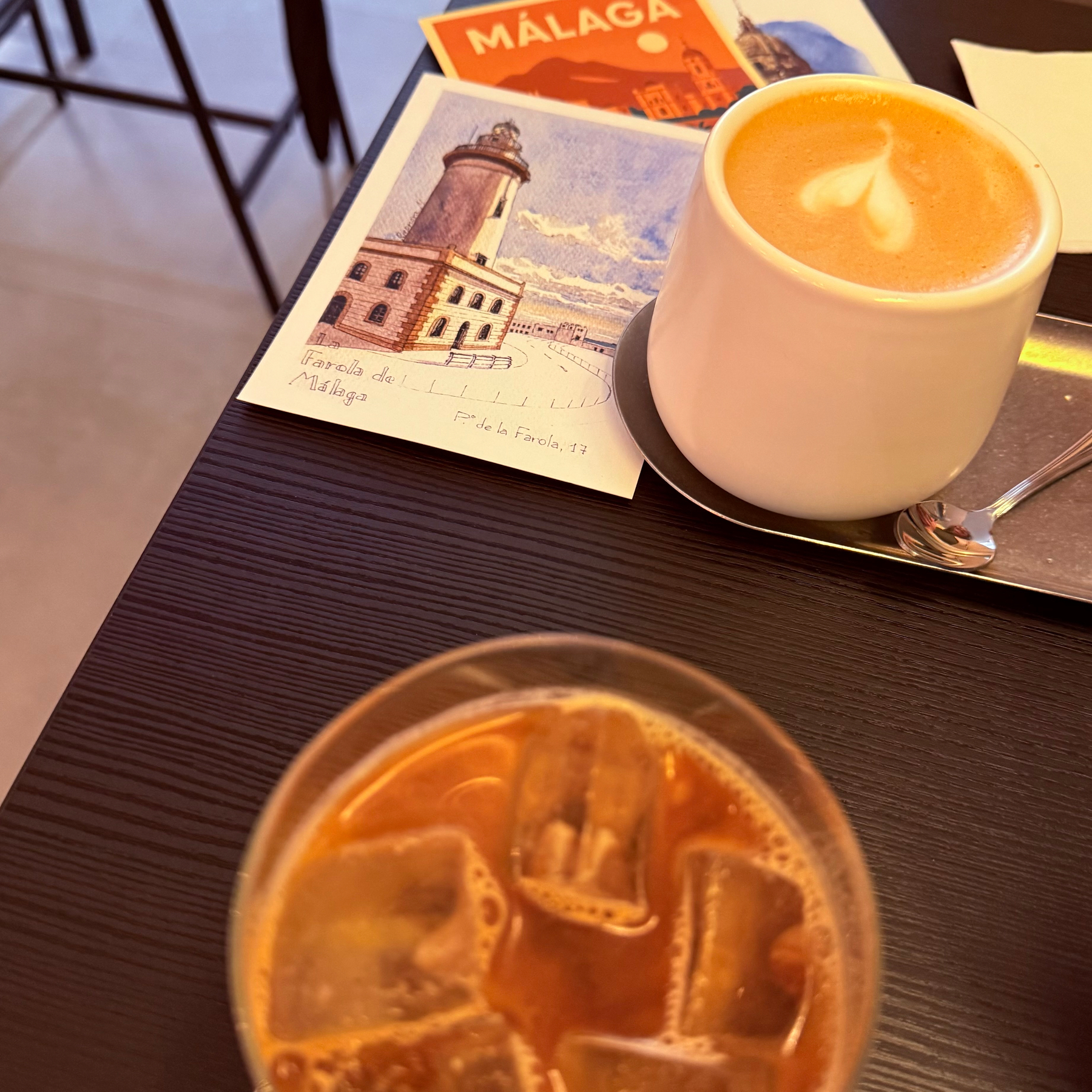 A table features a cup of latte art and an iced drink, with postcards of Málaga nearby.