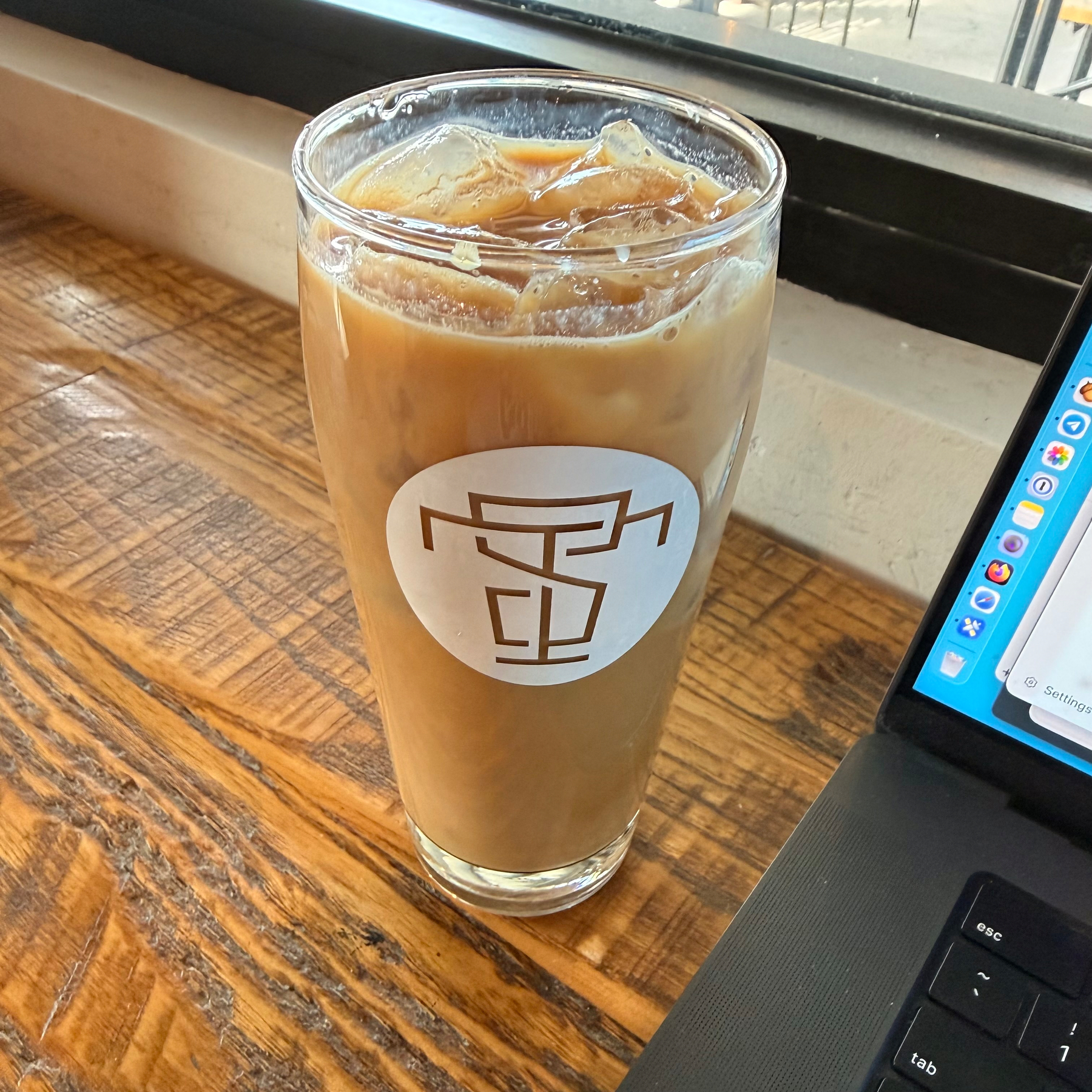 A glass of iced coffee is placed on a wooden table next to an open laptop.