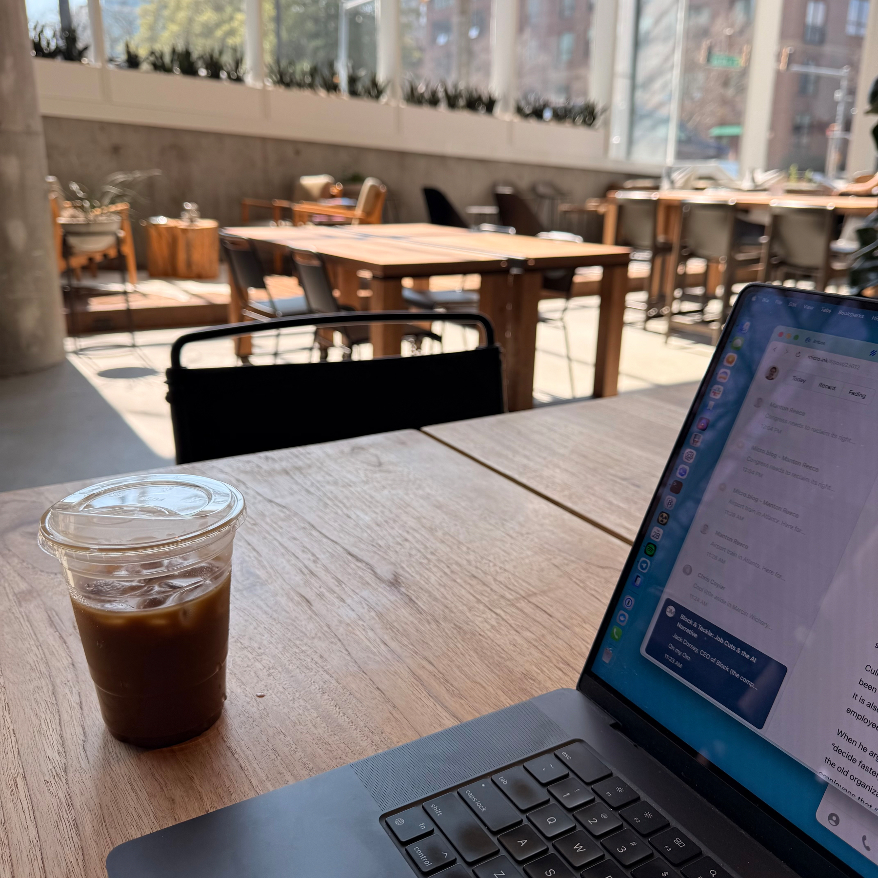 A laptop and an iced coffee sit on a wooden table in a well-lit cafe with a modern interior.